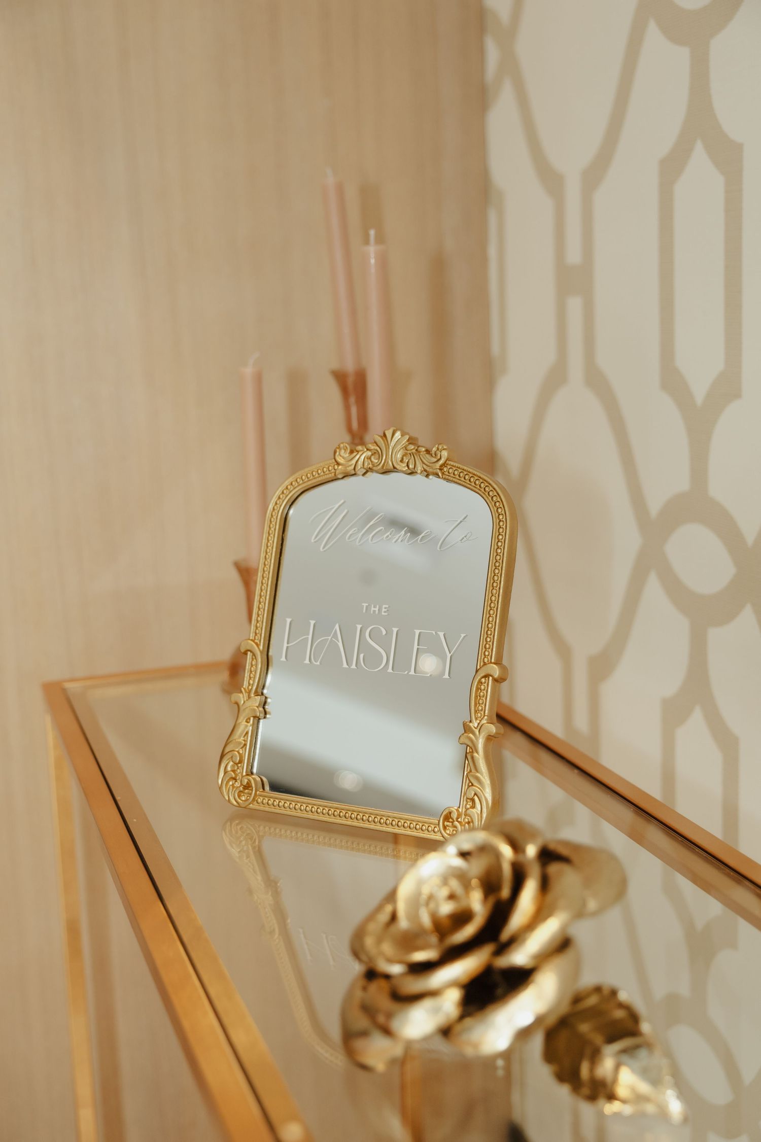Elegant gold-framed mirror with ornate details mounted on beige wallpaper with geometric pattern.