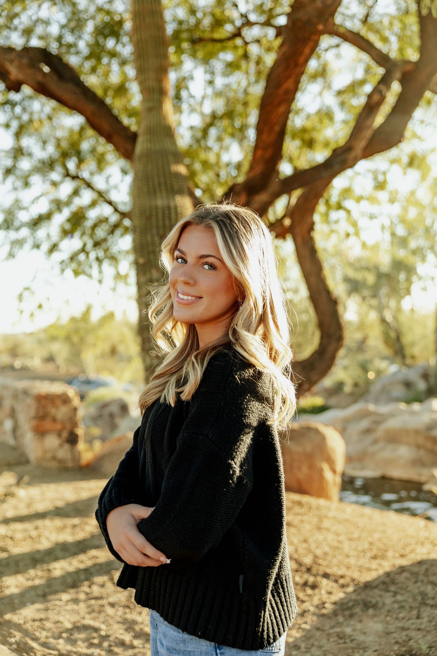 Woman in black sweater poses outdoors against sunlit trees and natural backdrop in autumn setting.