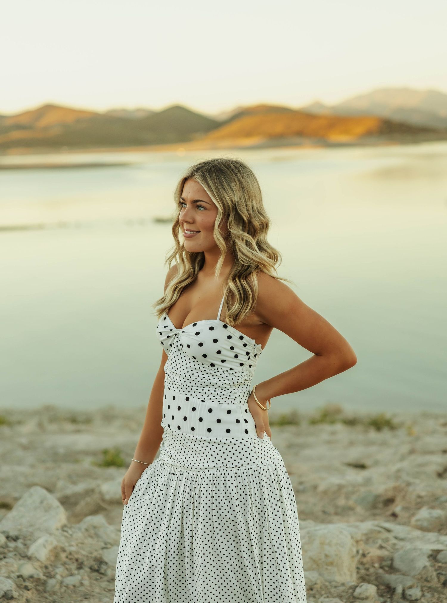 A white polka dot sundress with ruffle details is modeled against a scenic desert mountain landscape at sunset.