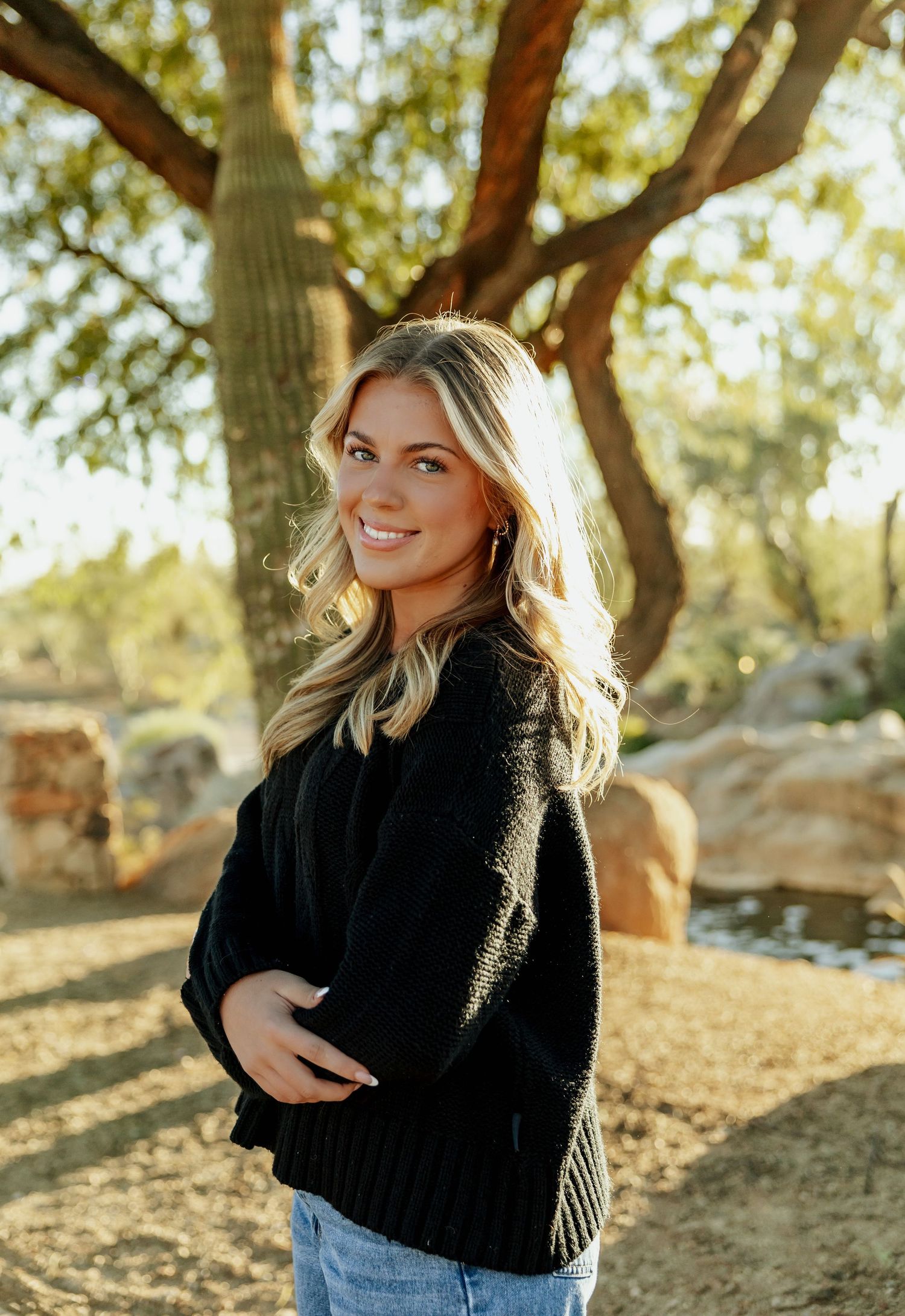 Person in black sweater and jeans poses outdoors in autumn sunlight with trees in background.