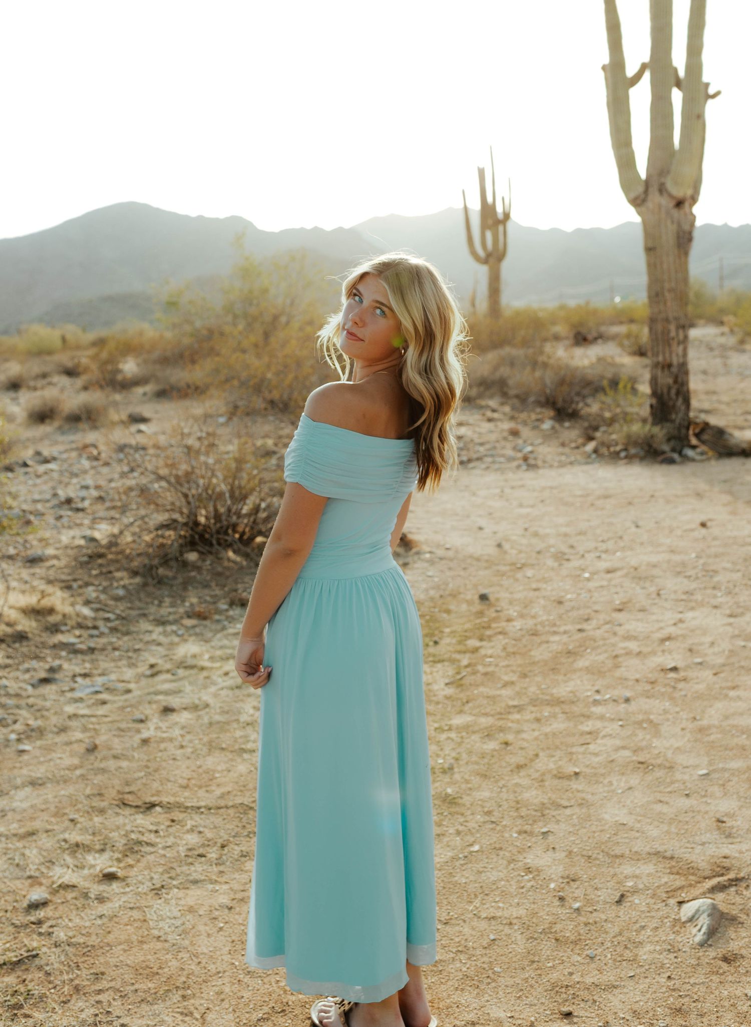 Woman in mint off-shoulder midi dress poses in desert landscape with saguaro cactus and mountain backdrop.