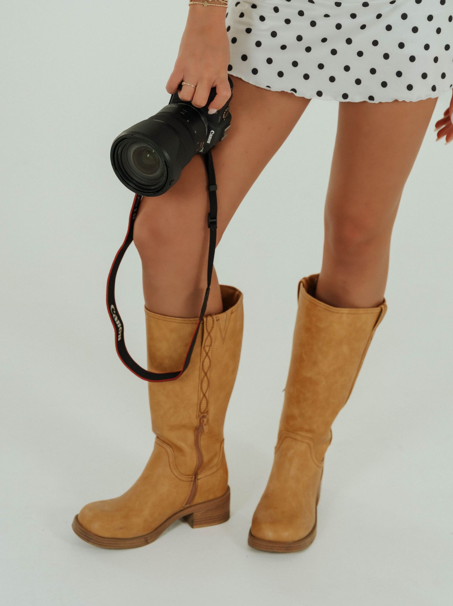 A person wearing a polka dot dress holds a black camera while wearing tan leather knee-high boots against a white background.