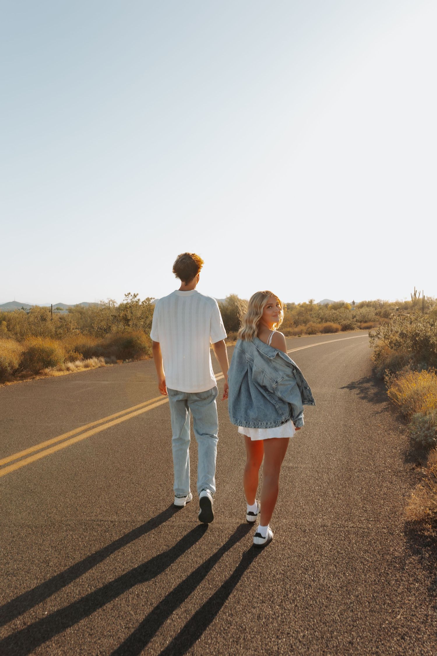 Two people walking down an empty desert road at sunset casting long shadows on the pavement.