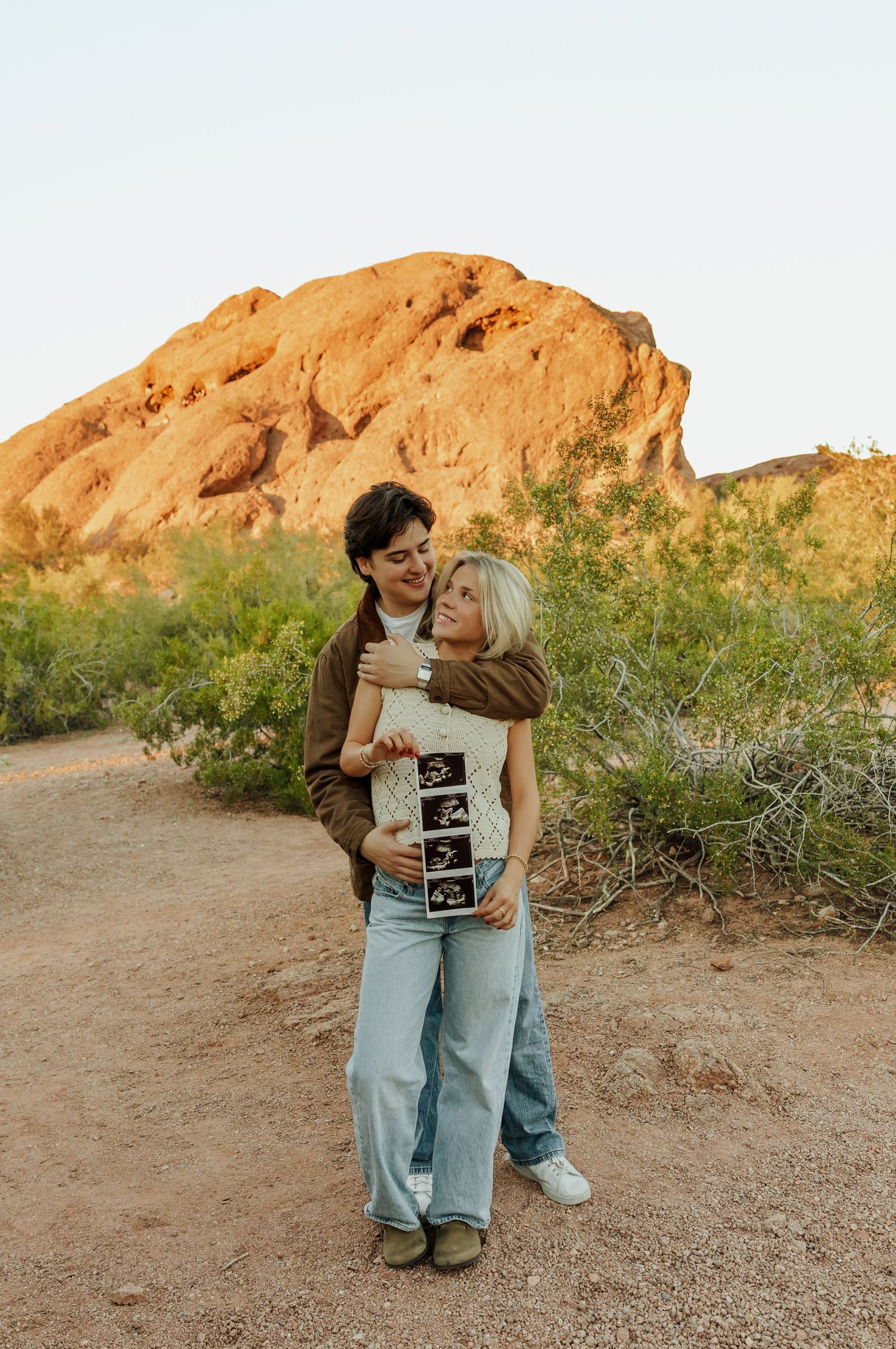 A couple shares a tender moment while holding an ultrasound photo against a desert mountain backdrop at sunset.