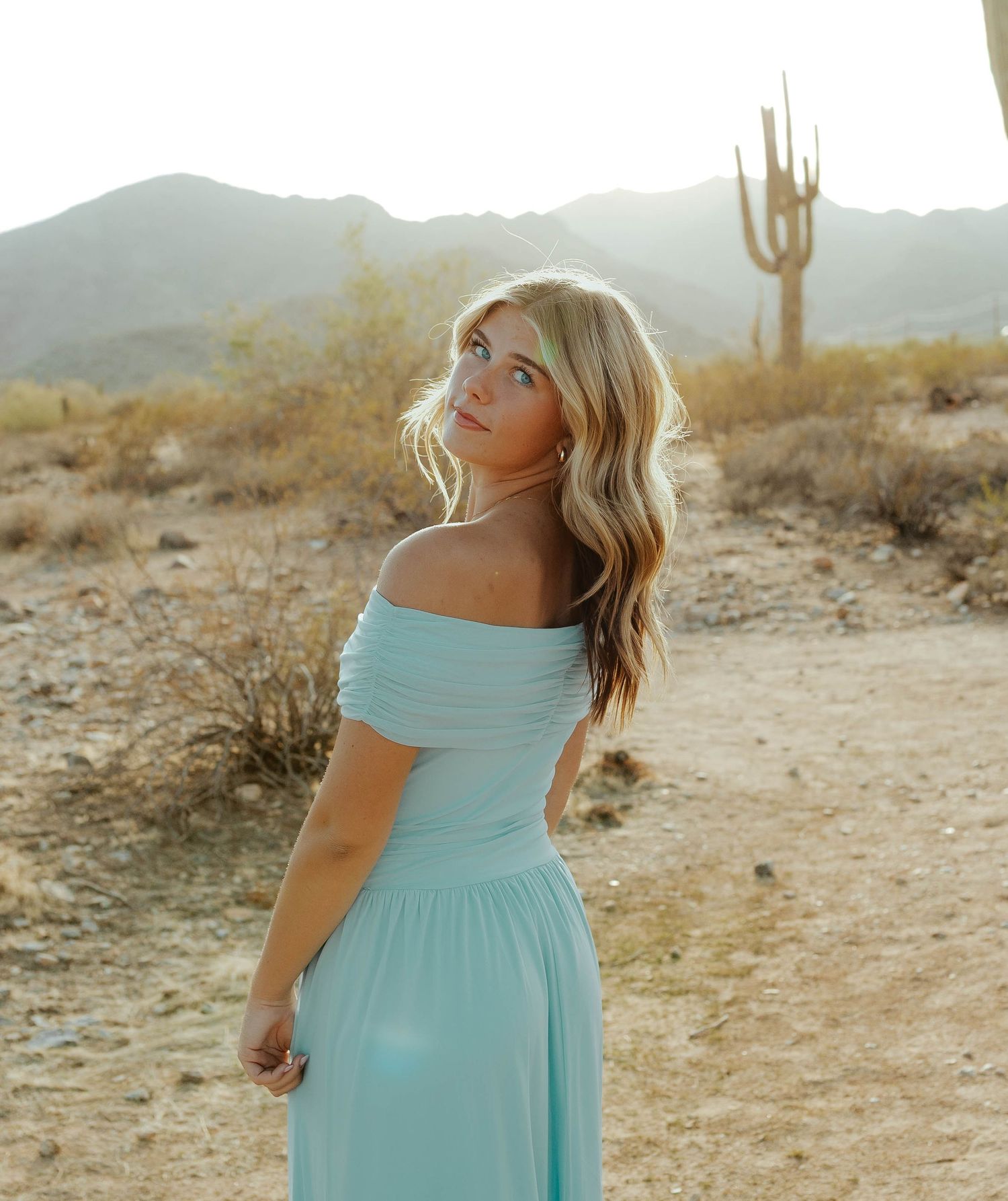 A light blue dress flows in the desert breeze during a senior grad sunset photoshoot with cactus and mountains.