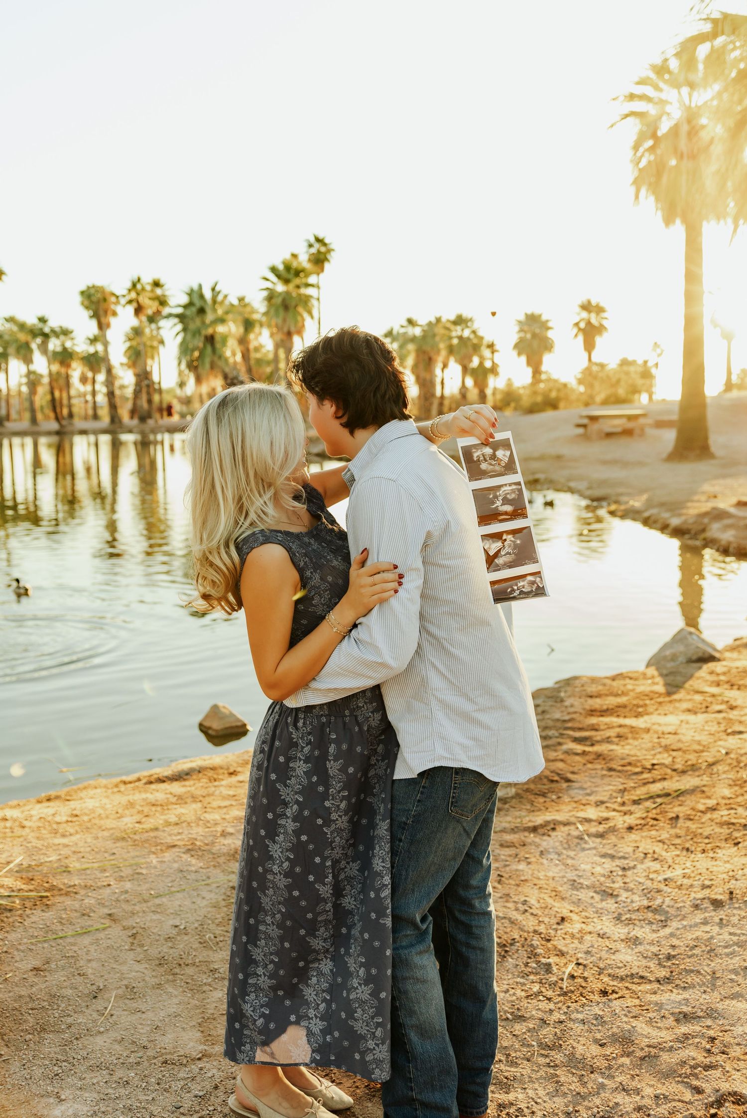 A romantic sunset moment by a palm-lined lake as a couple embraces while holding ultrasound photos.