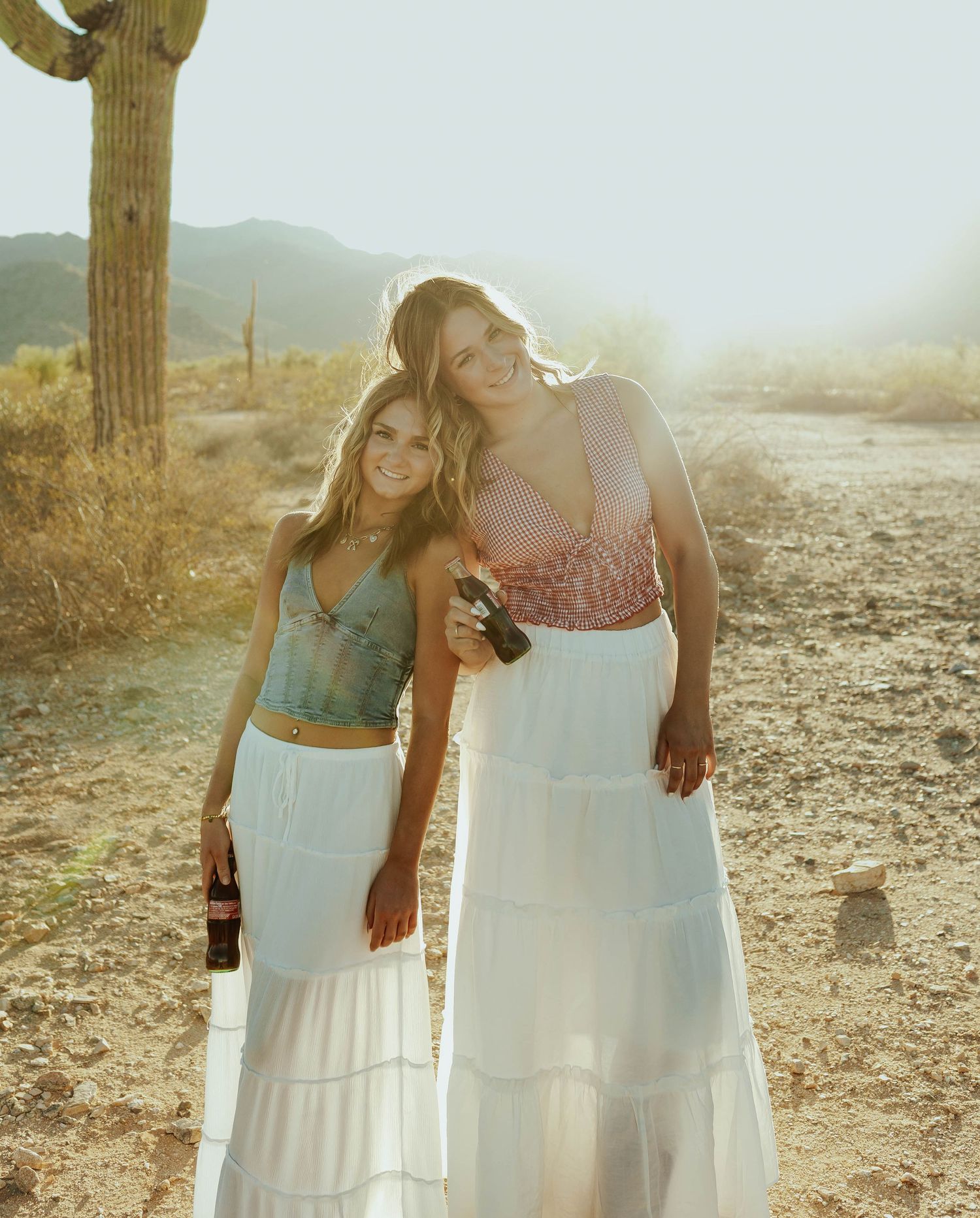 Friends in white dresses pose together in a desert landscape with saguaro cacti at sunset.