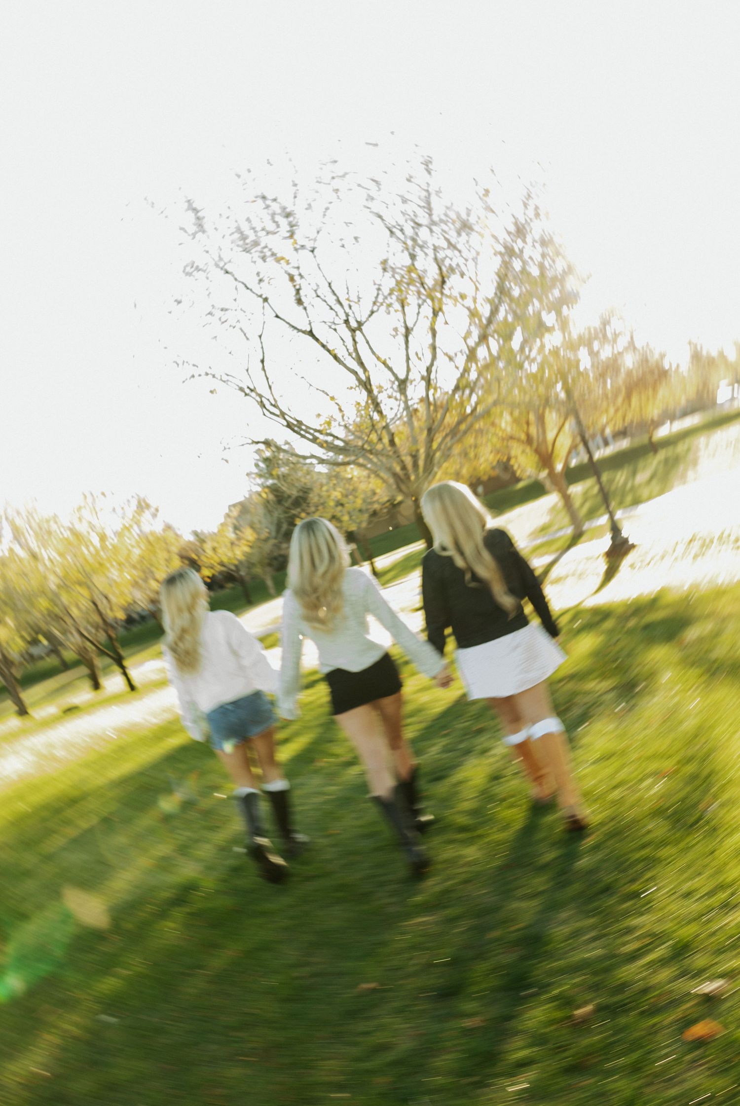 Three blonde girls holding hands running through a lush green park on a sunny autumn day.