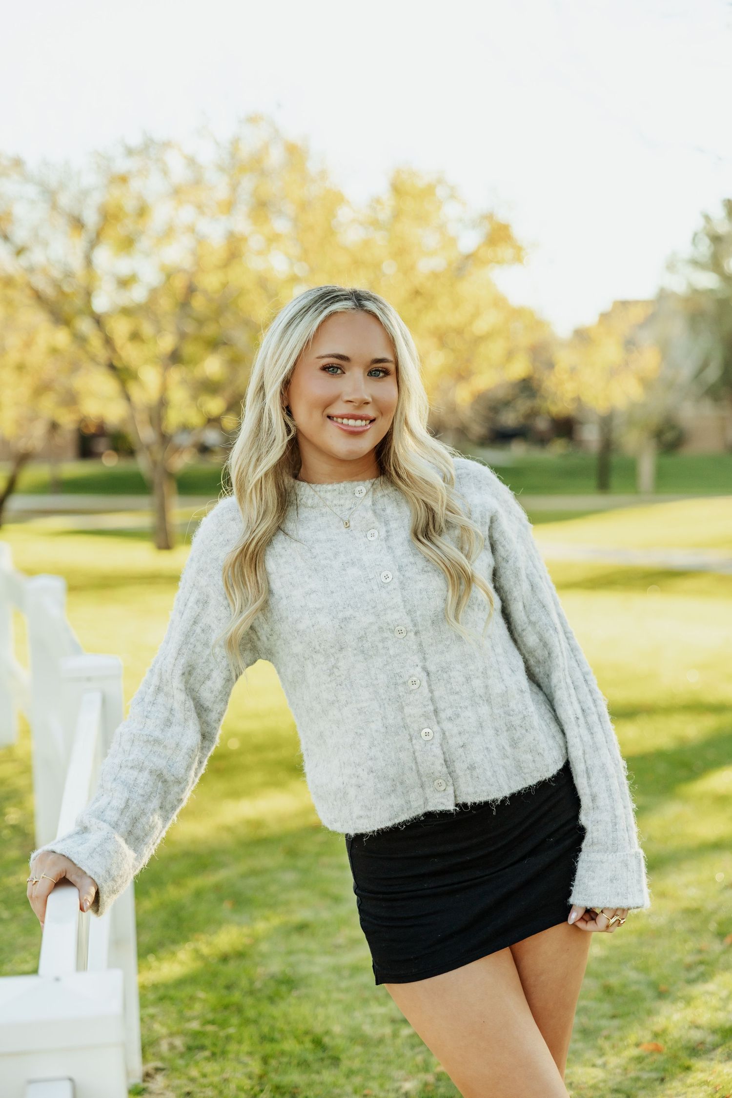 Smiling blonde woman in gray sweater and black mini skirt posing outdoors in a lush green park setting.