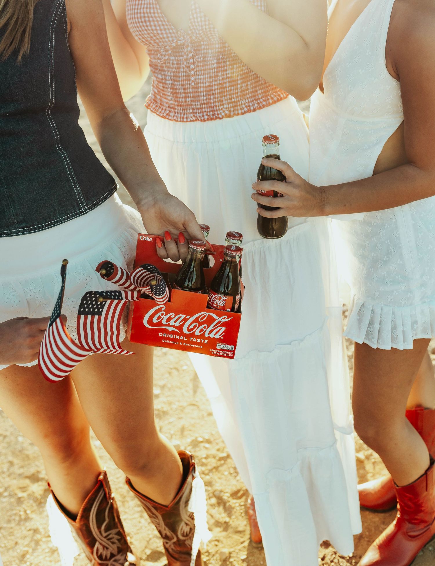 People in summer outfits holding glass Coca-Cola bottles with red and white striped straws at an outdoor gathering.