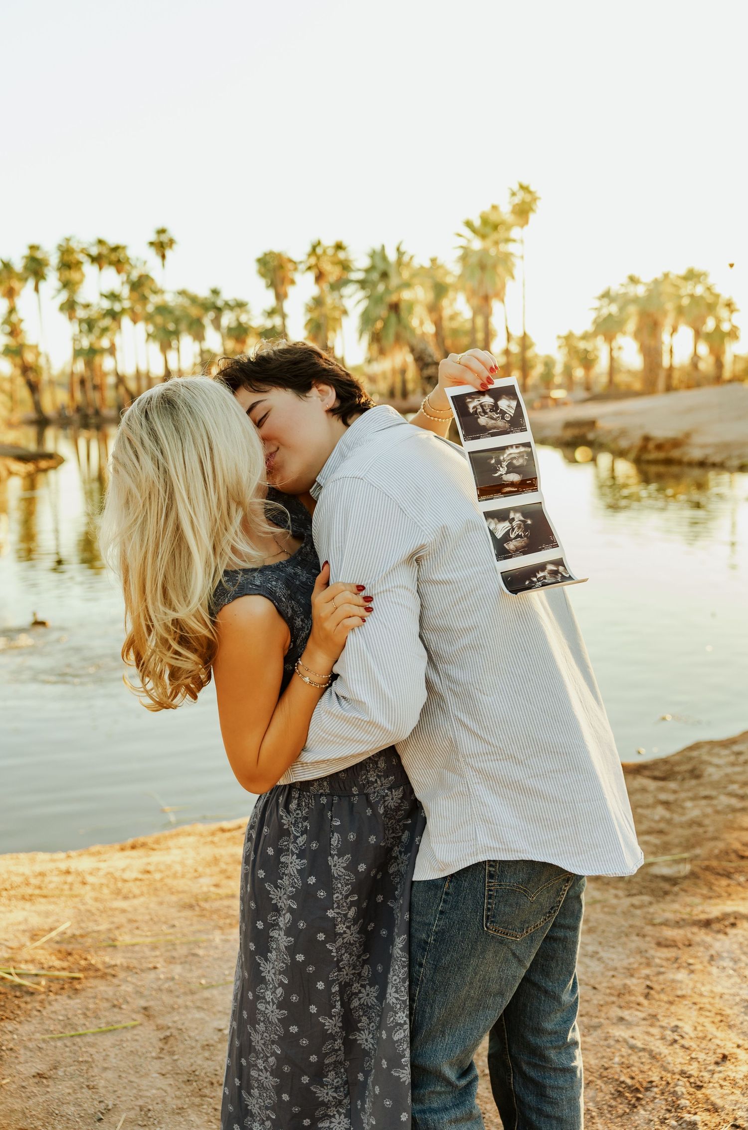 A couple shares a romantic moment on a beach with palm trees and water at sunset while holding ultrasound photos.