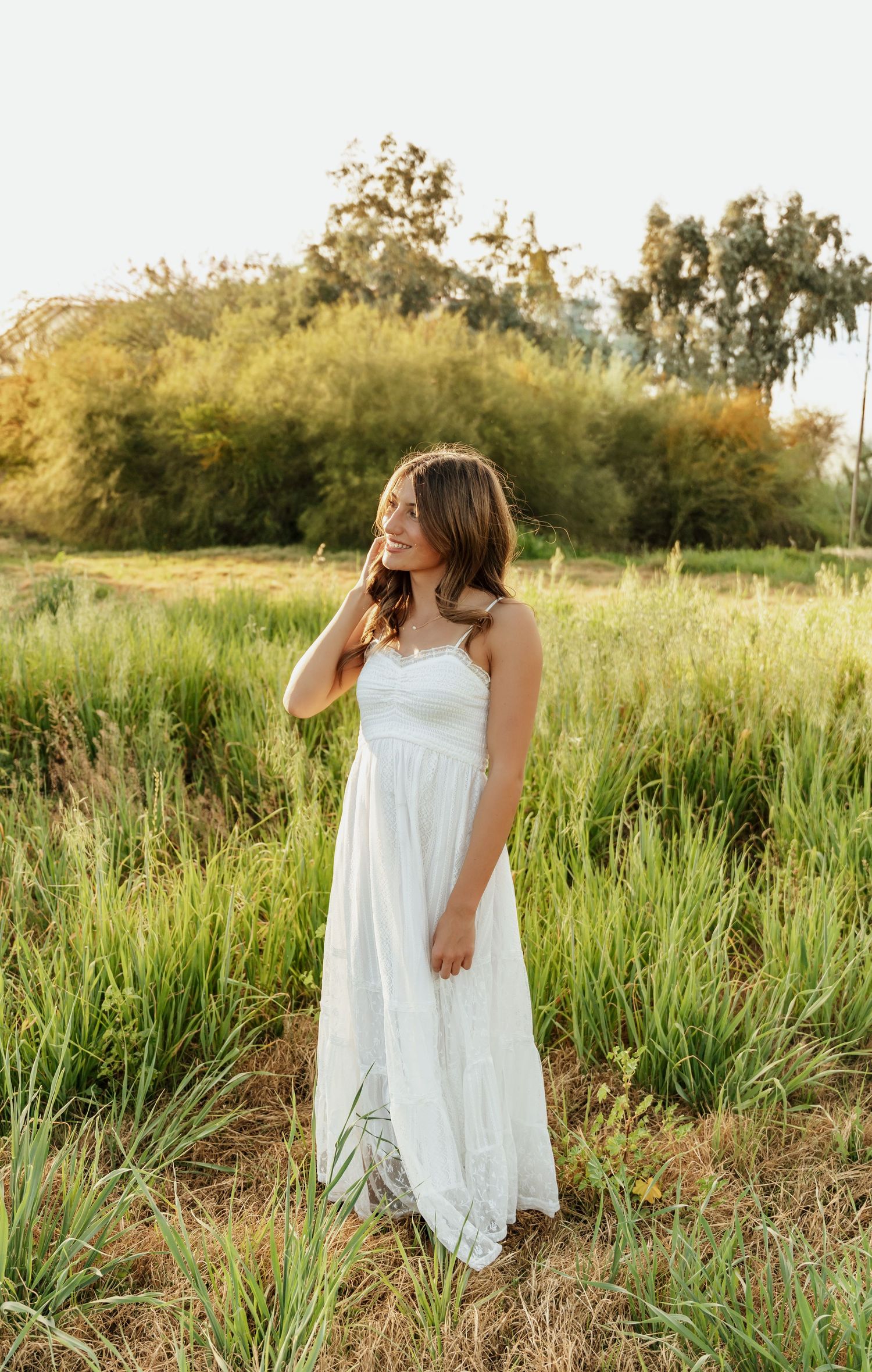 A figure in a flowing white dress stands gracefully among tall grass and wildflowers at golden hour.