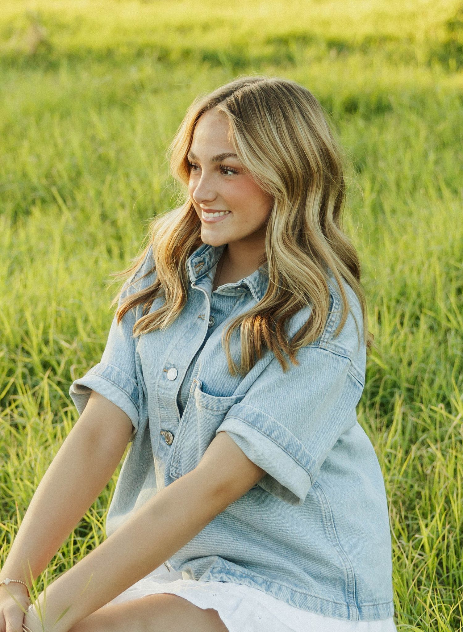 Woman in light denim shirt smiling while sitting in grassy field.
