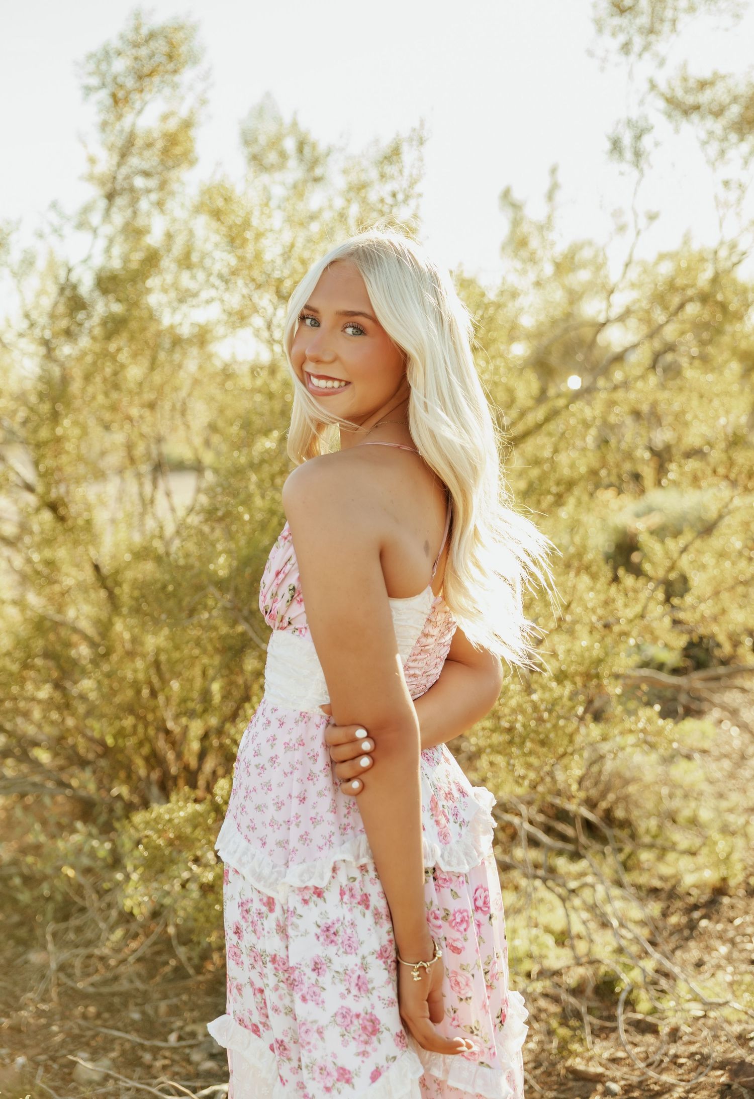 A person in a floral summer dress poses against a sunlit natural backdrop with desert foliage.