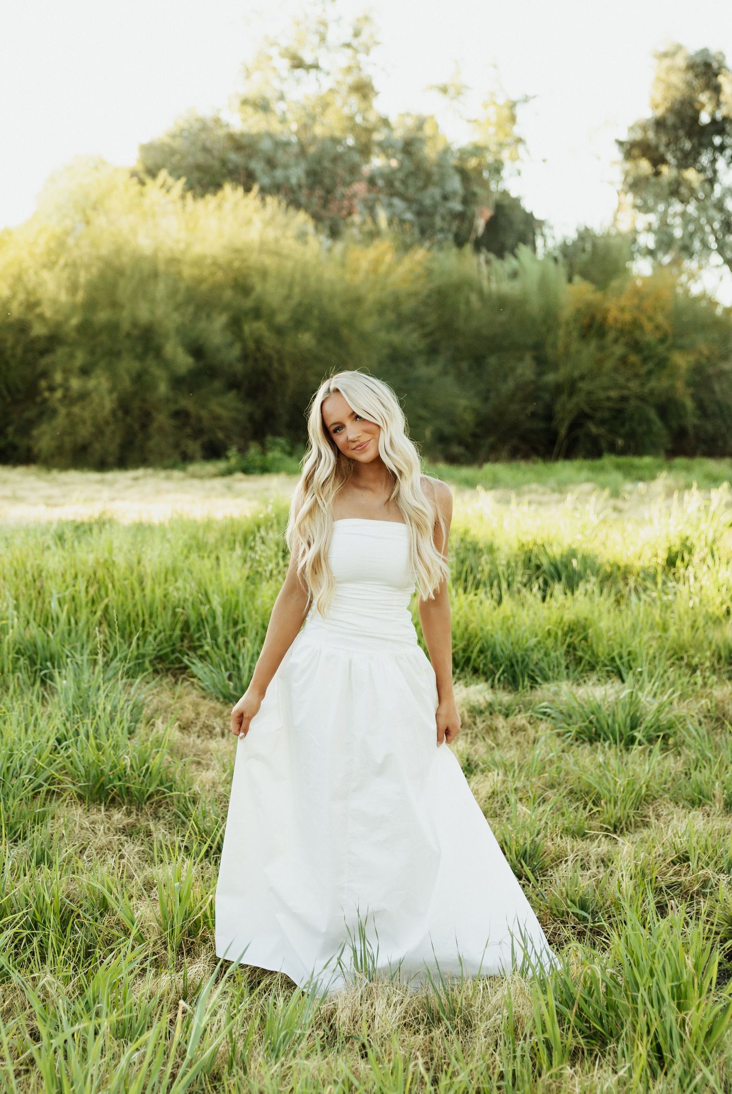 A person in a flowing white dress walks through a sunlit meadow of tall grass during golden hour.
