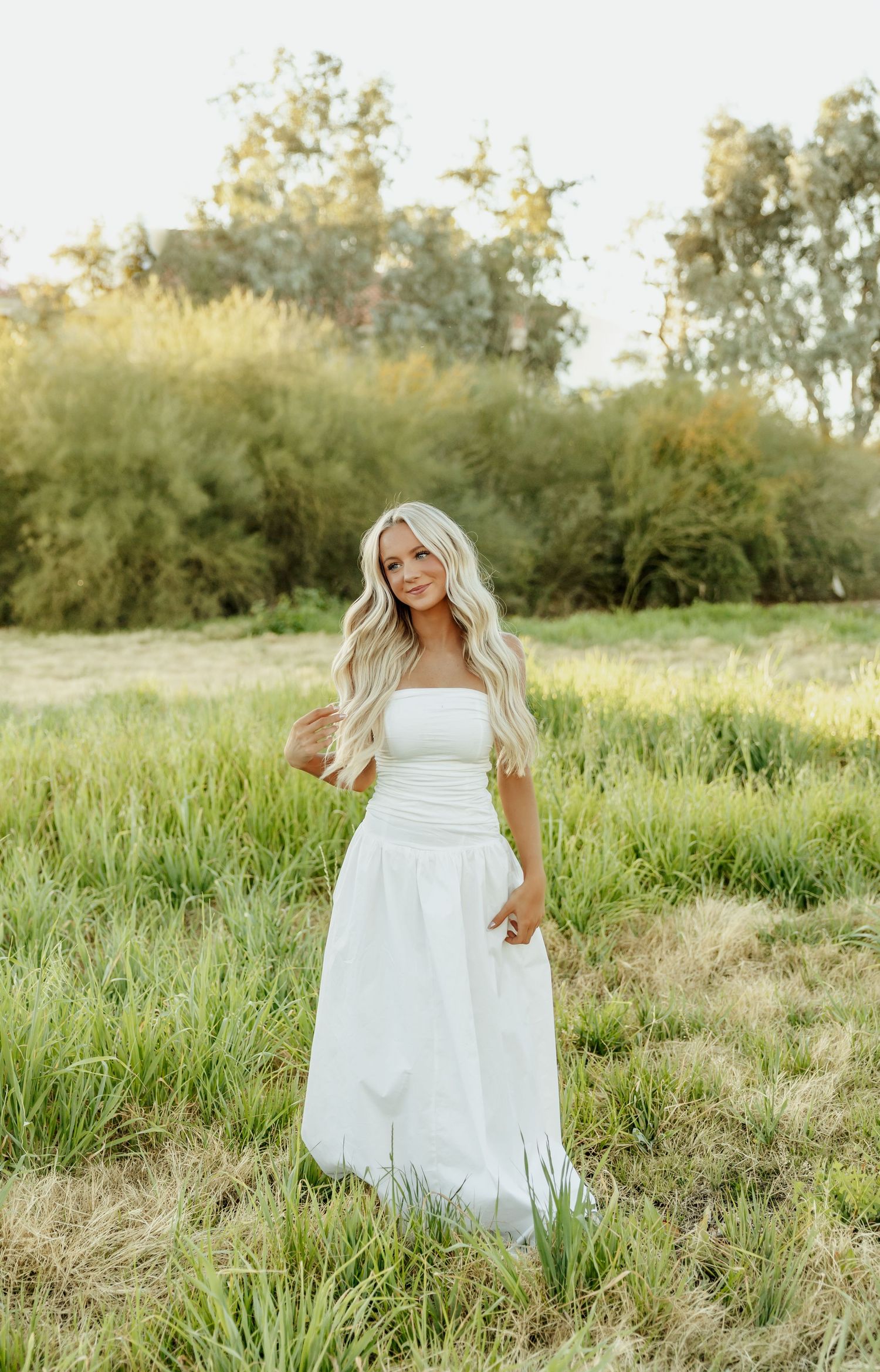 A person in a flowing white dress poses gracefully in a sunlit meadow with tall grass and trees in the background.