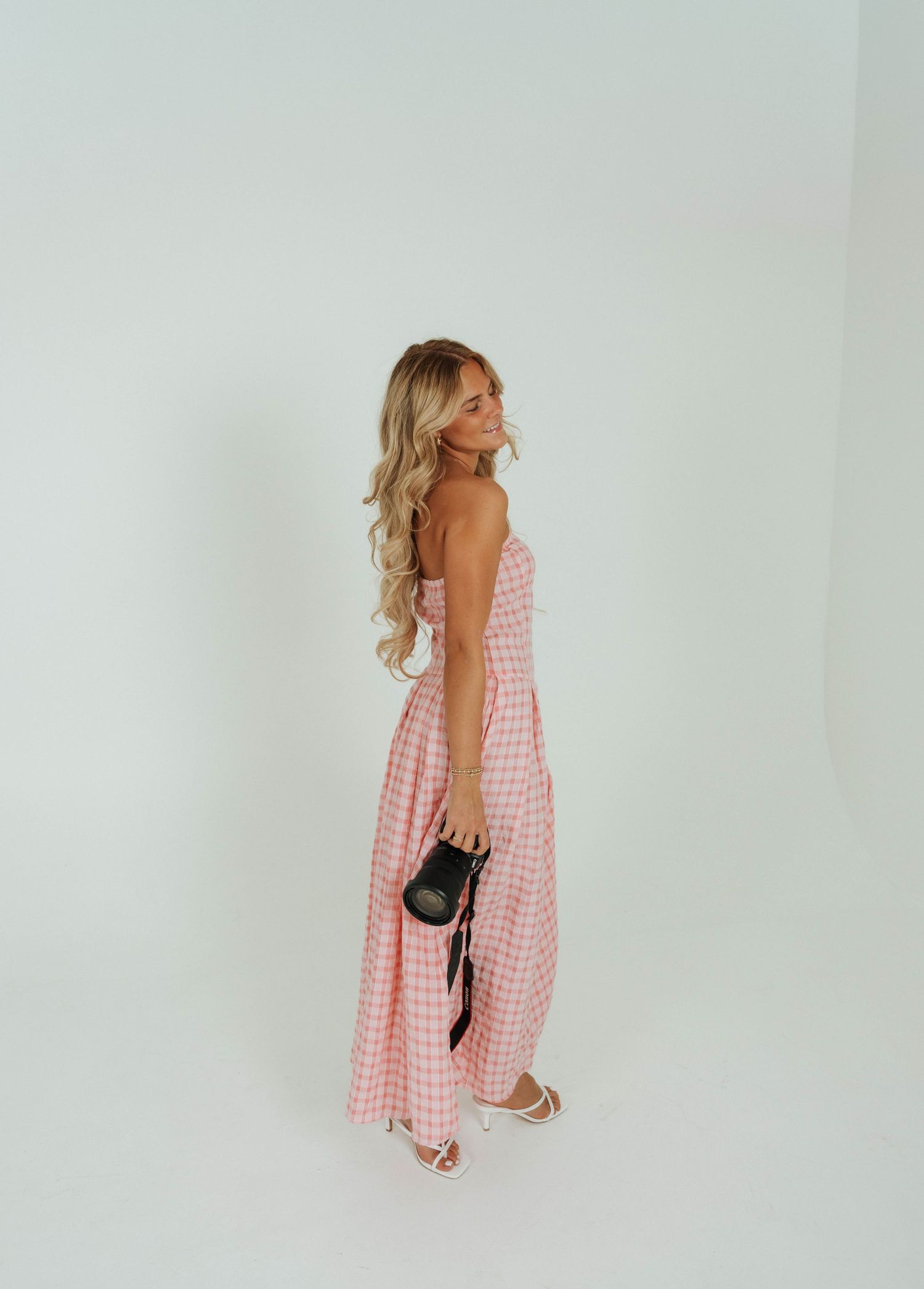 Fashion poses in a pink gingham summer dress holding vintage camera against white backdrop.
