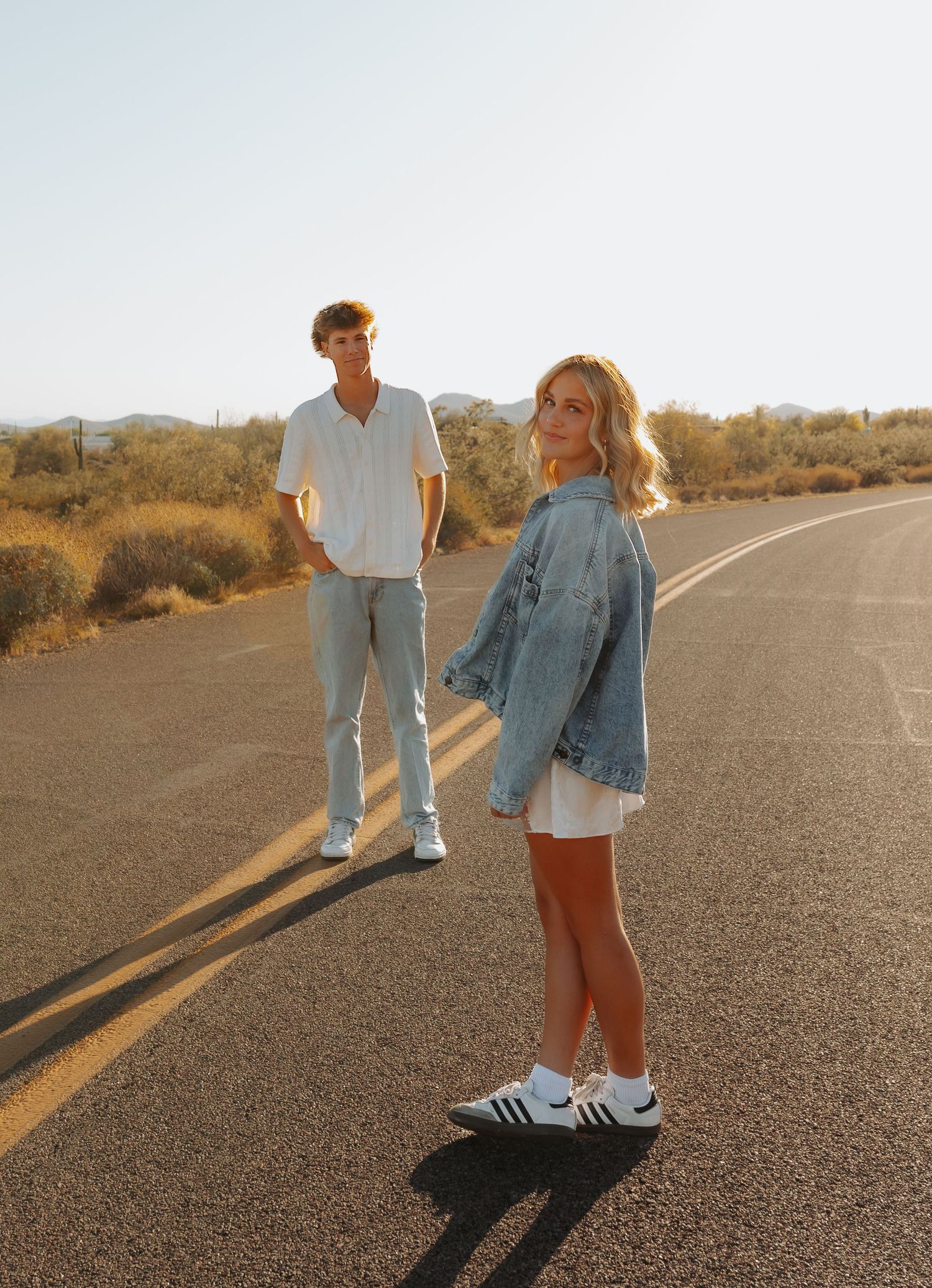Couple in denim outfits walking on empty desert road at sunset casting long shadows on the pavement.