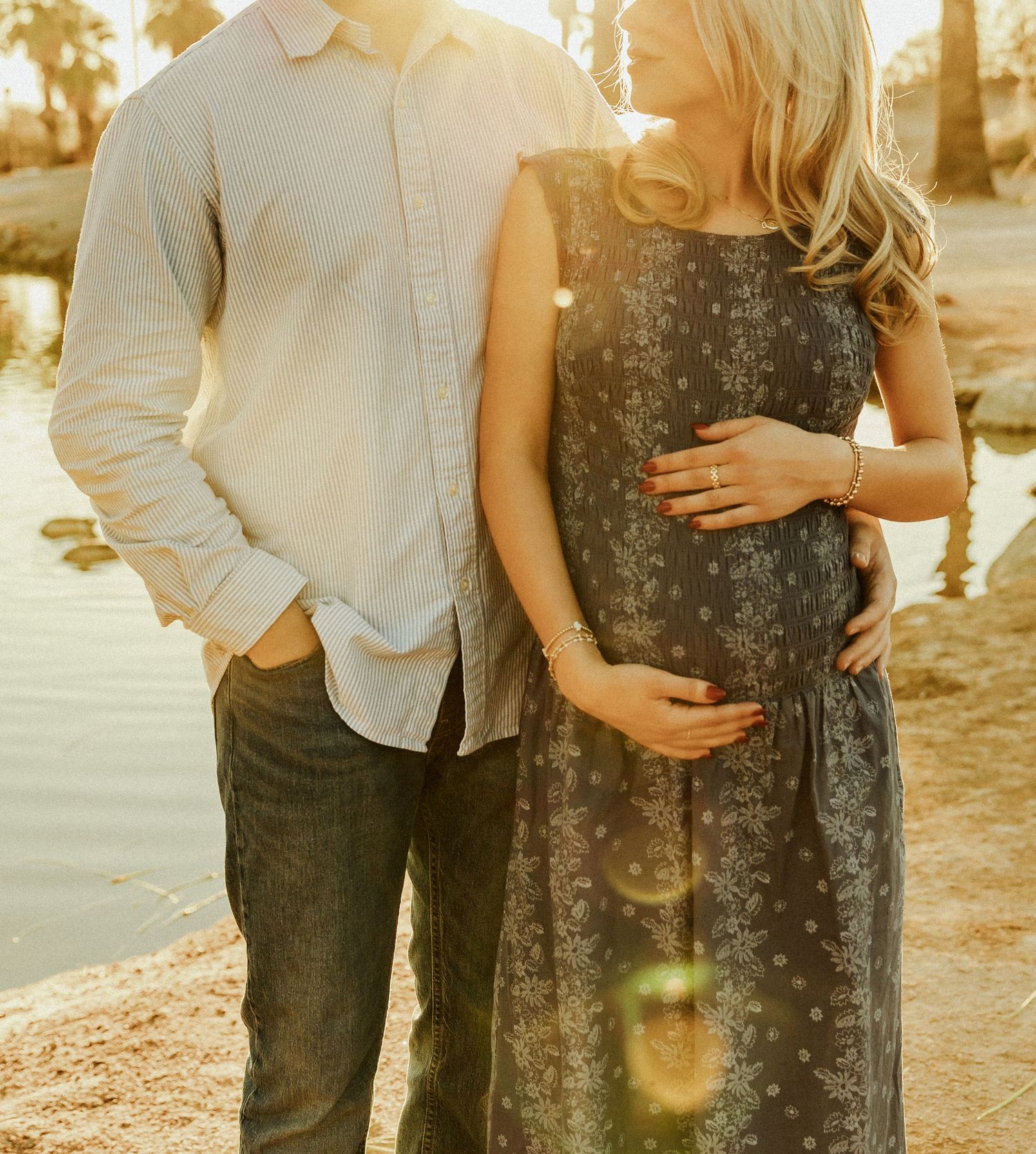 A couple stands together in warm sunset light, with the woman wearing a floral dress and cradling her pregnant belly.