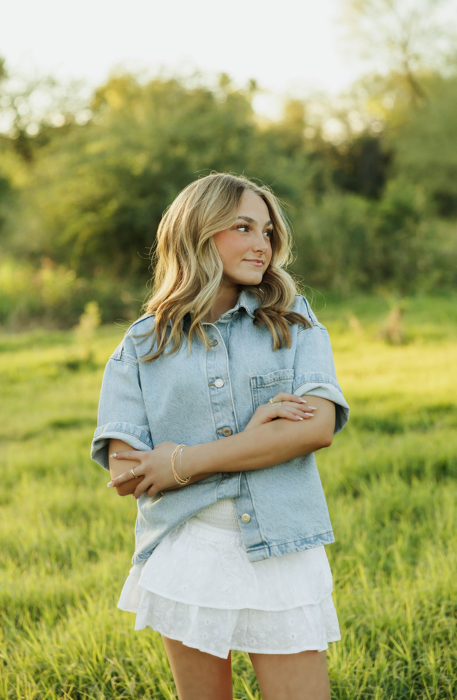 Woman in denim jacket poses confidently against green natural backdrop.