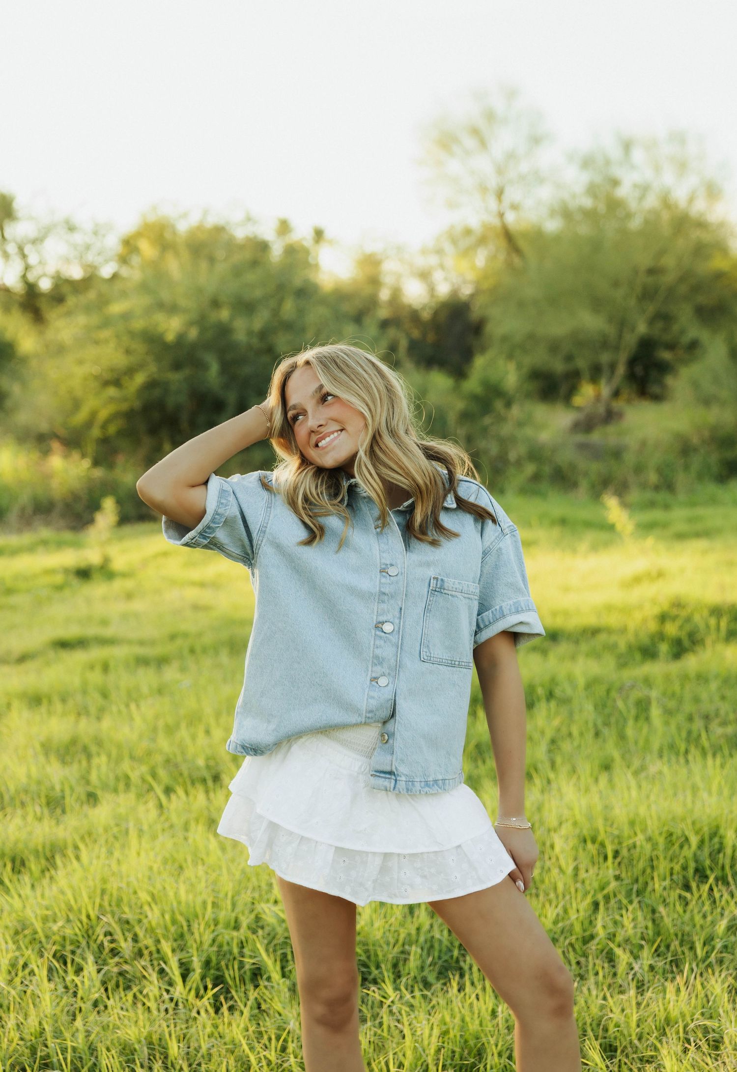 Woman in denim shirt and white skirt smiling in sunlit meadow.