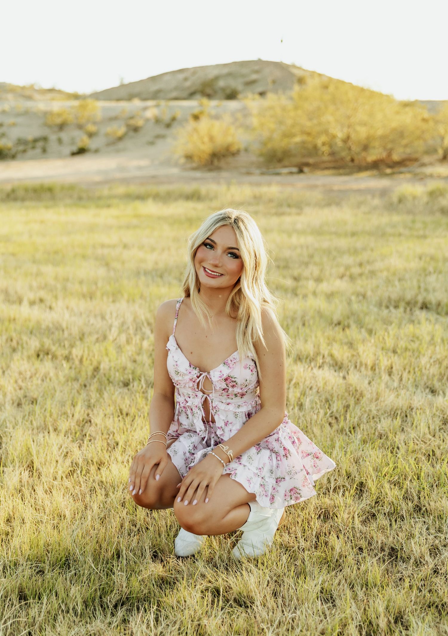 Smiling blonde woman in floral mini dress crouching in a golden grass field with sandy hills in the background.