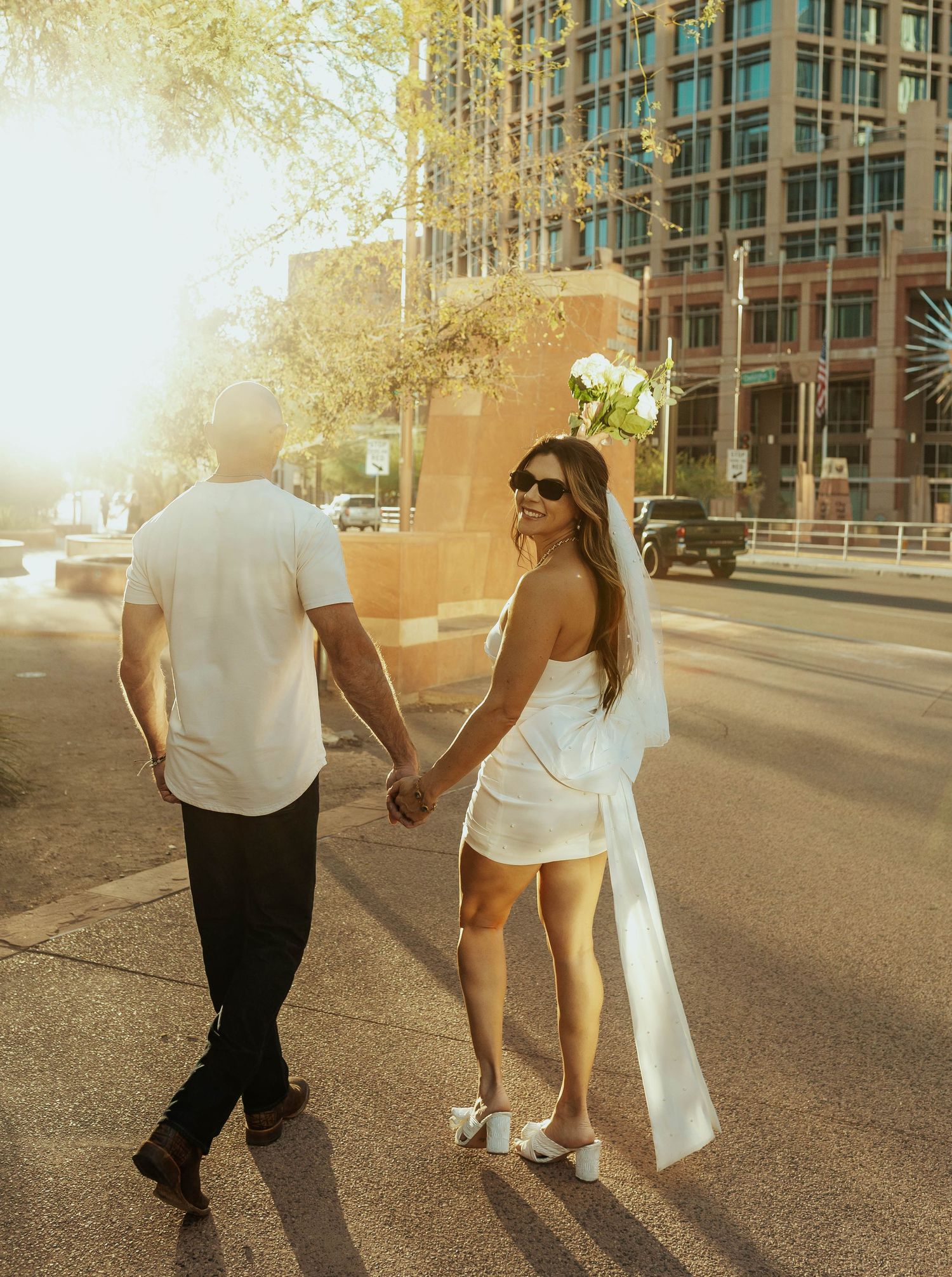 Couple holding hands while walking down a city street with tall buildings and trees. Elopement Photographer