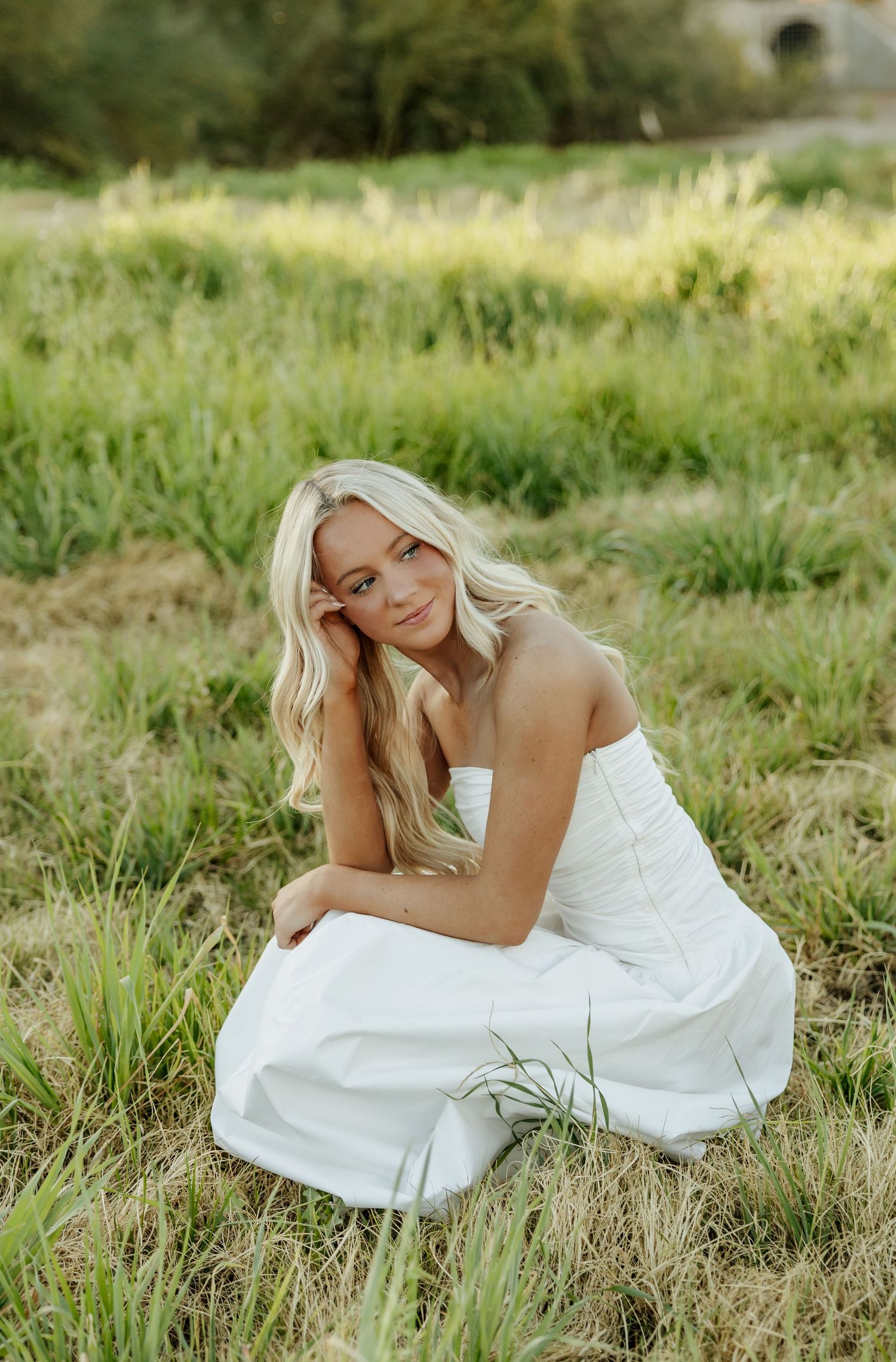 A person in a flowing white dress reclines gracefully in tall grass during a sunny outdoor photo session.