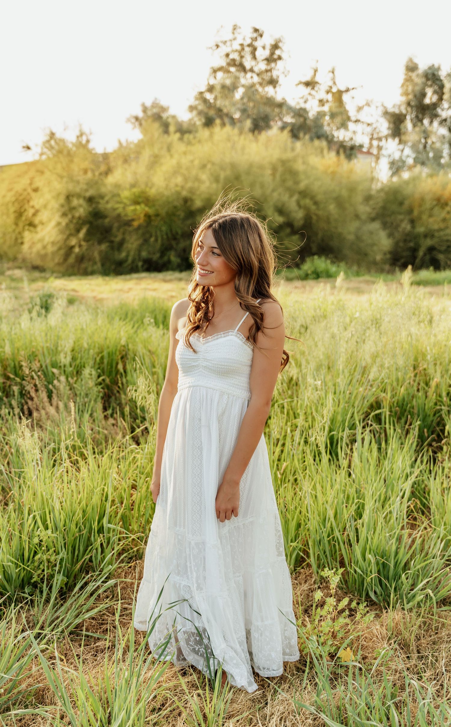 A person in a flowing white dress sits gracefully in tall summer grass during golden hour.