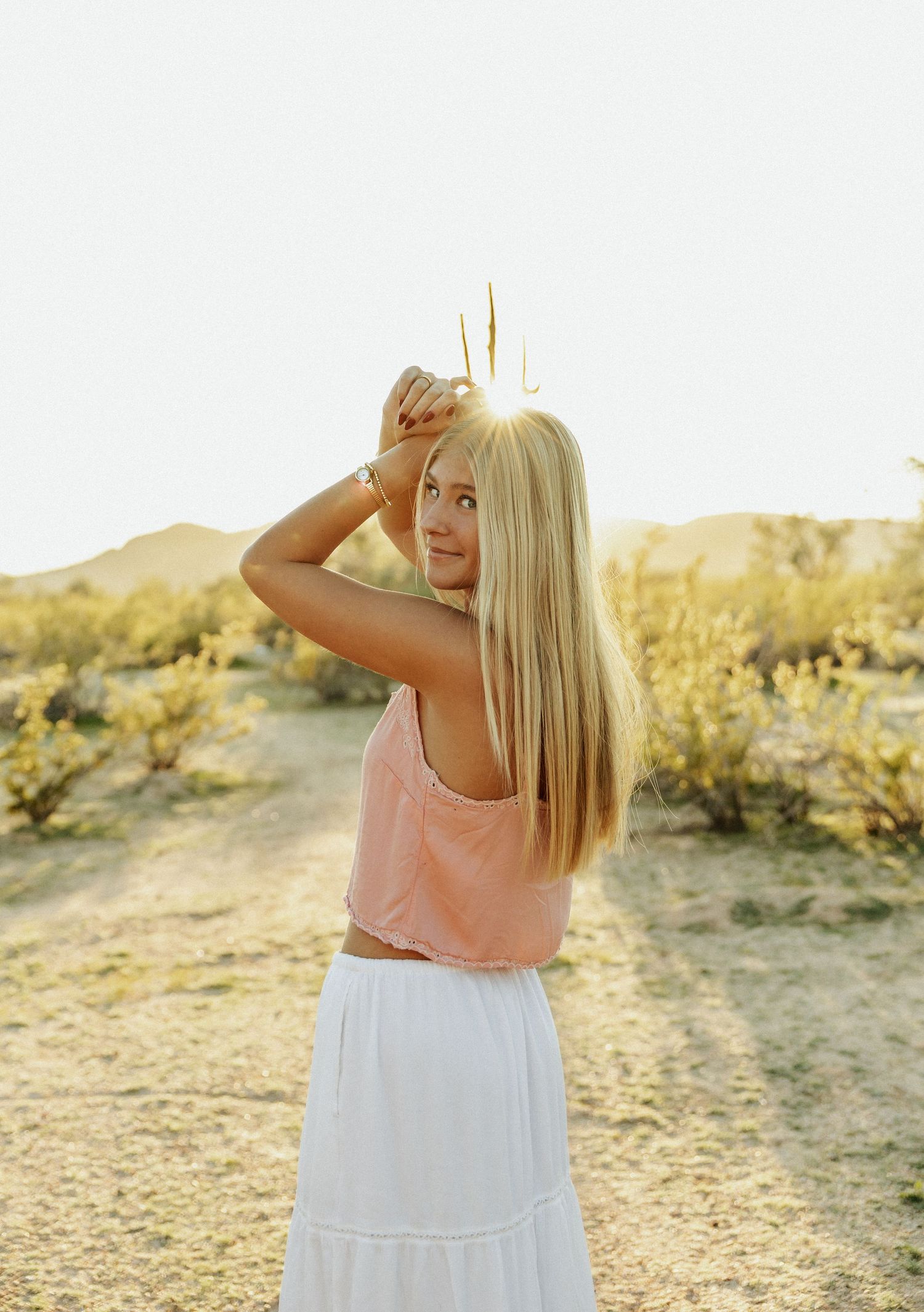 A person in a pink top and white skirt poses in a desert landscape during golden hour.