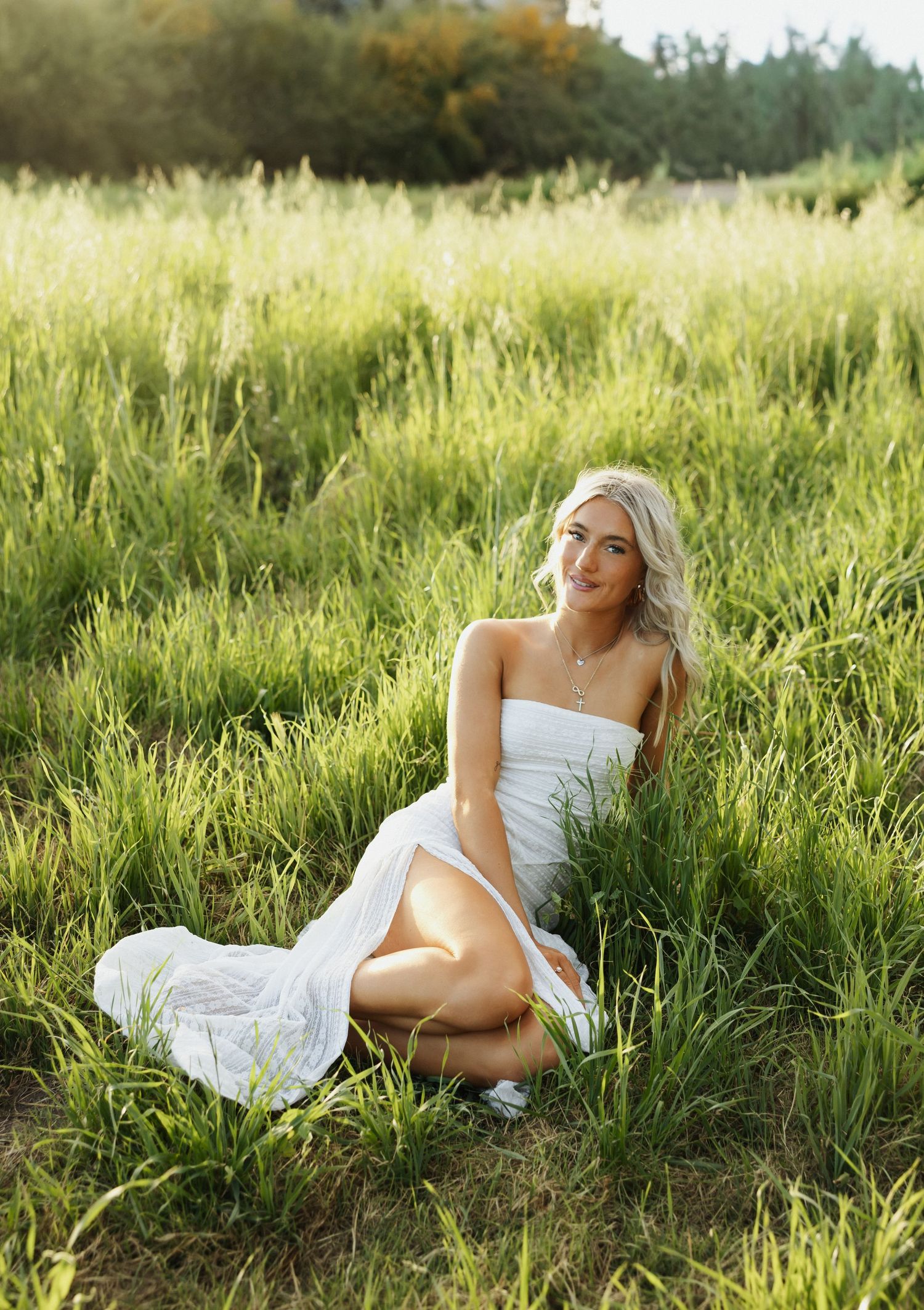 A figure in a white dress poses gracefully in tall summer grass during golden hour lighting.