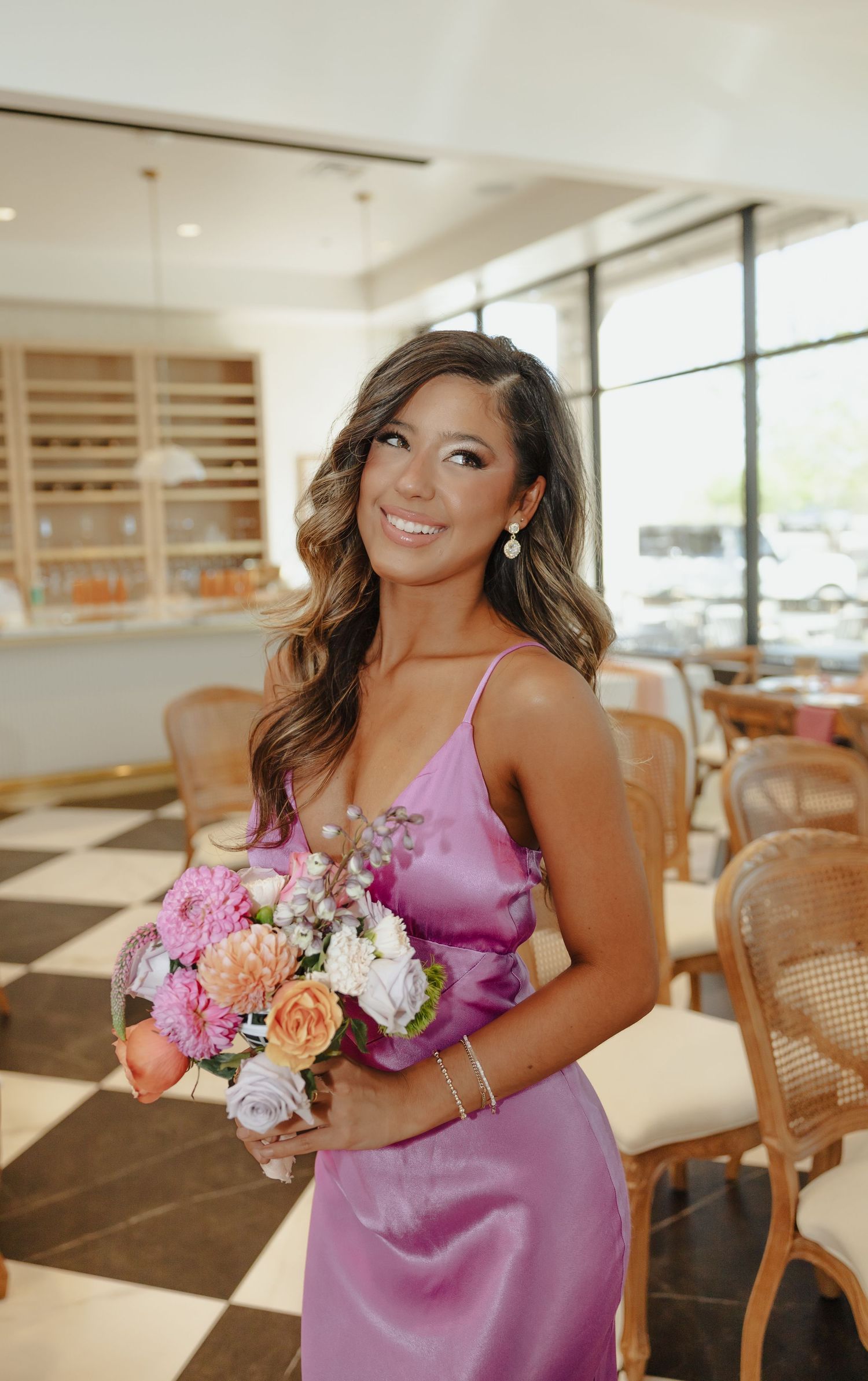Woman in pink silk dress holds colorful bouquet of flowers in a bright restaurant interior.
