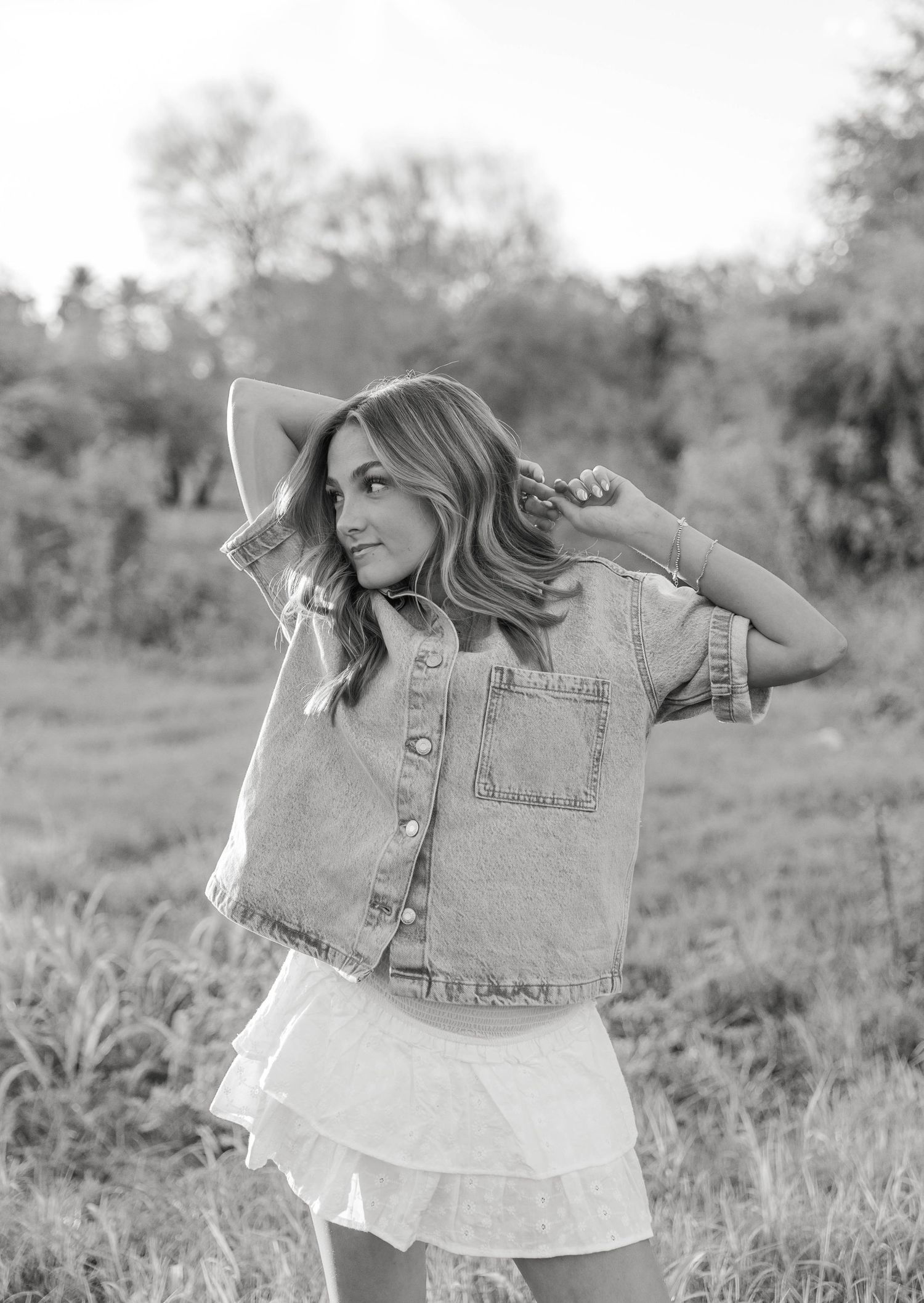 A black and white photo shows someone in a denim vest playfully posing in a grassy outdoor setting.