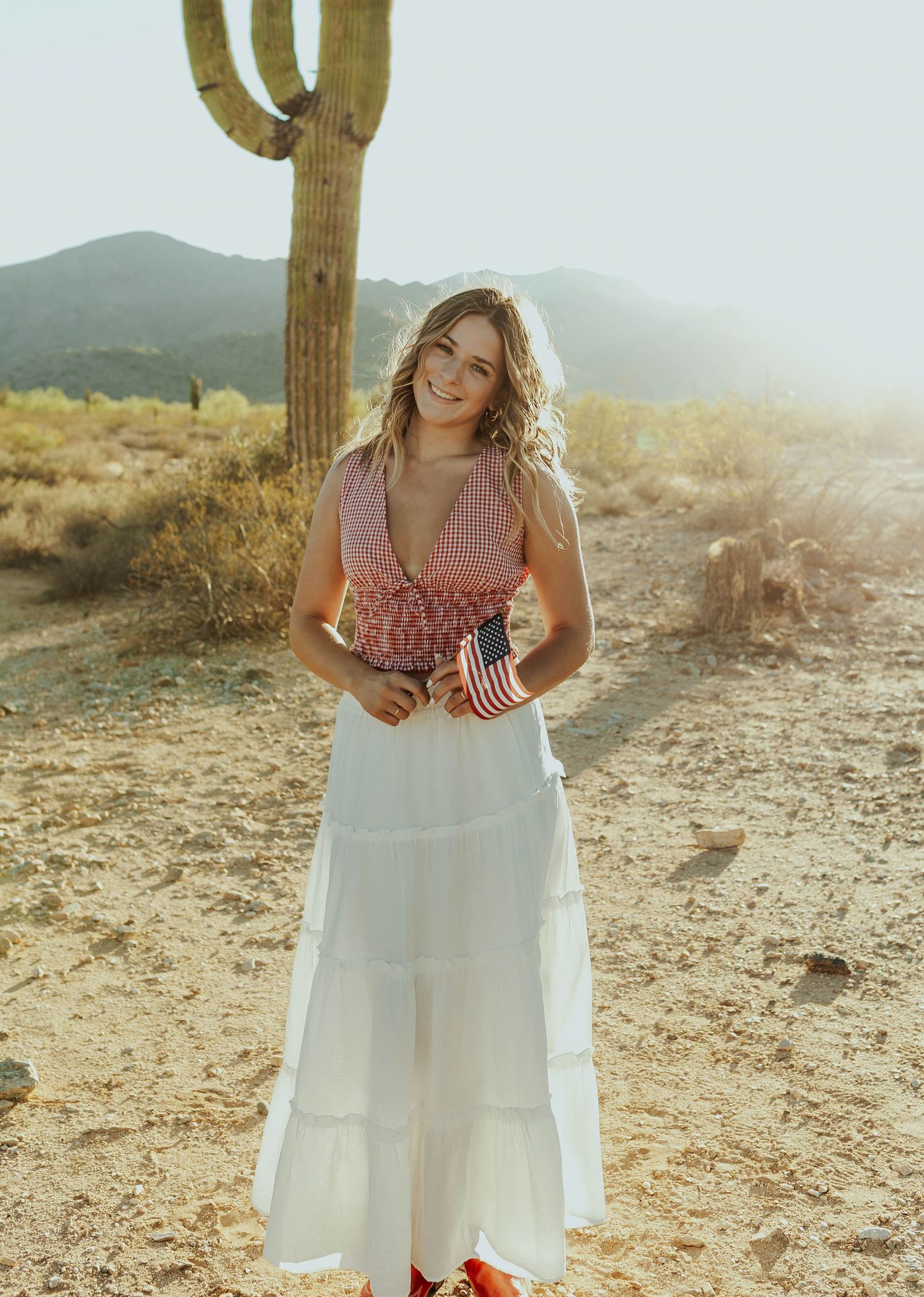 Woman in flowy white dress poses near desert cactus during sunset.