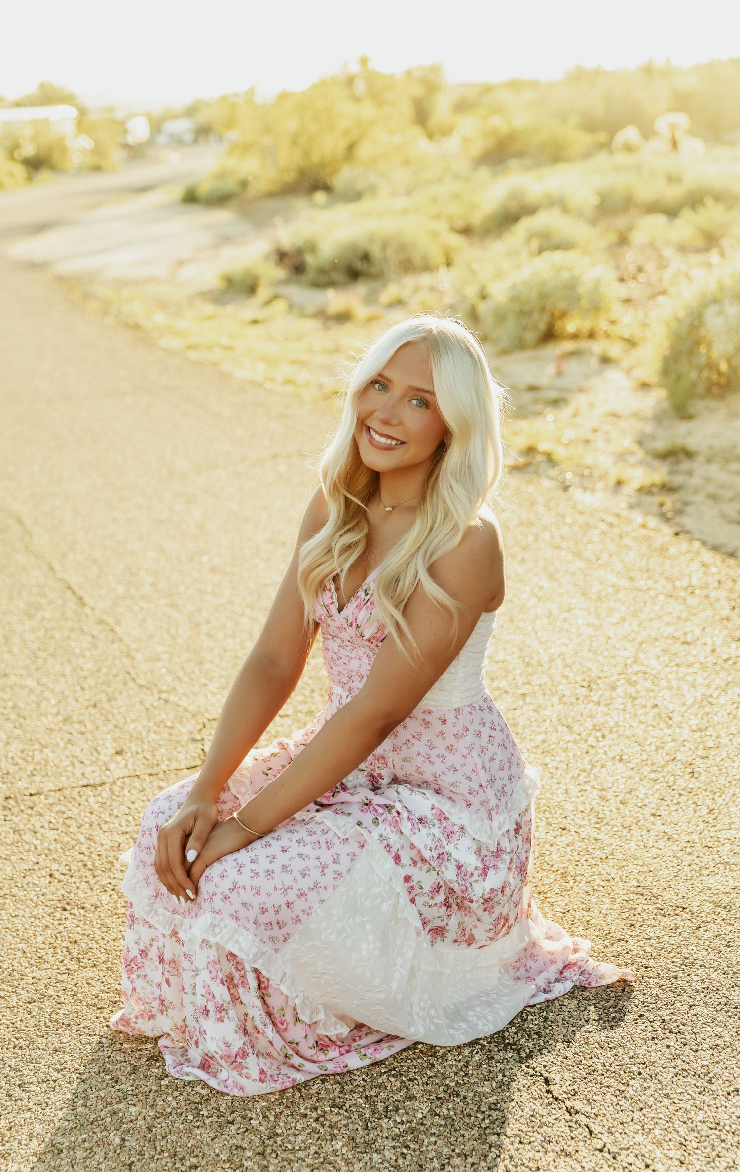 A woman in a white and pink floral dress sits on sandy ground during golden hour.
