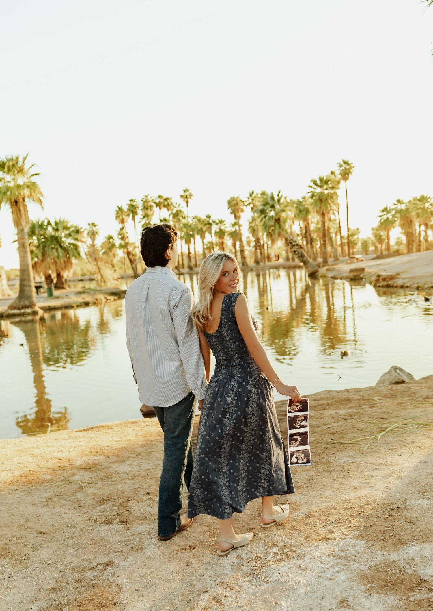 A couple walks along a sandy shoreline with palm trees and water reflections during golden hour sunset.