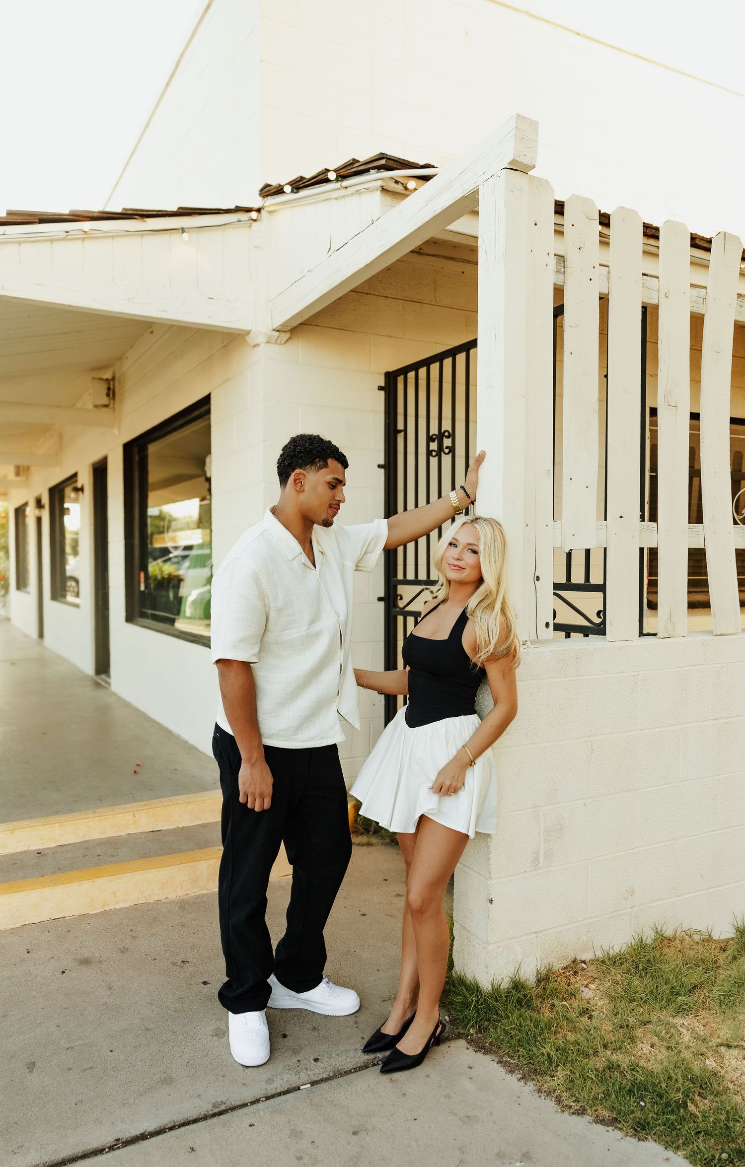 A man in white shirt and black pants stands with a blonde woman in black top and white skirt outside a building.