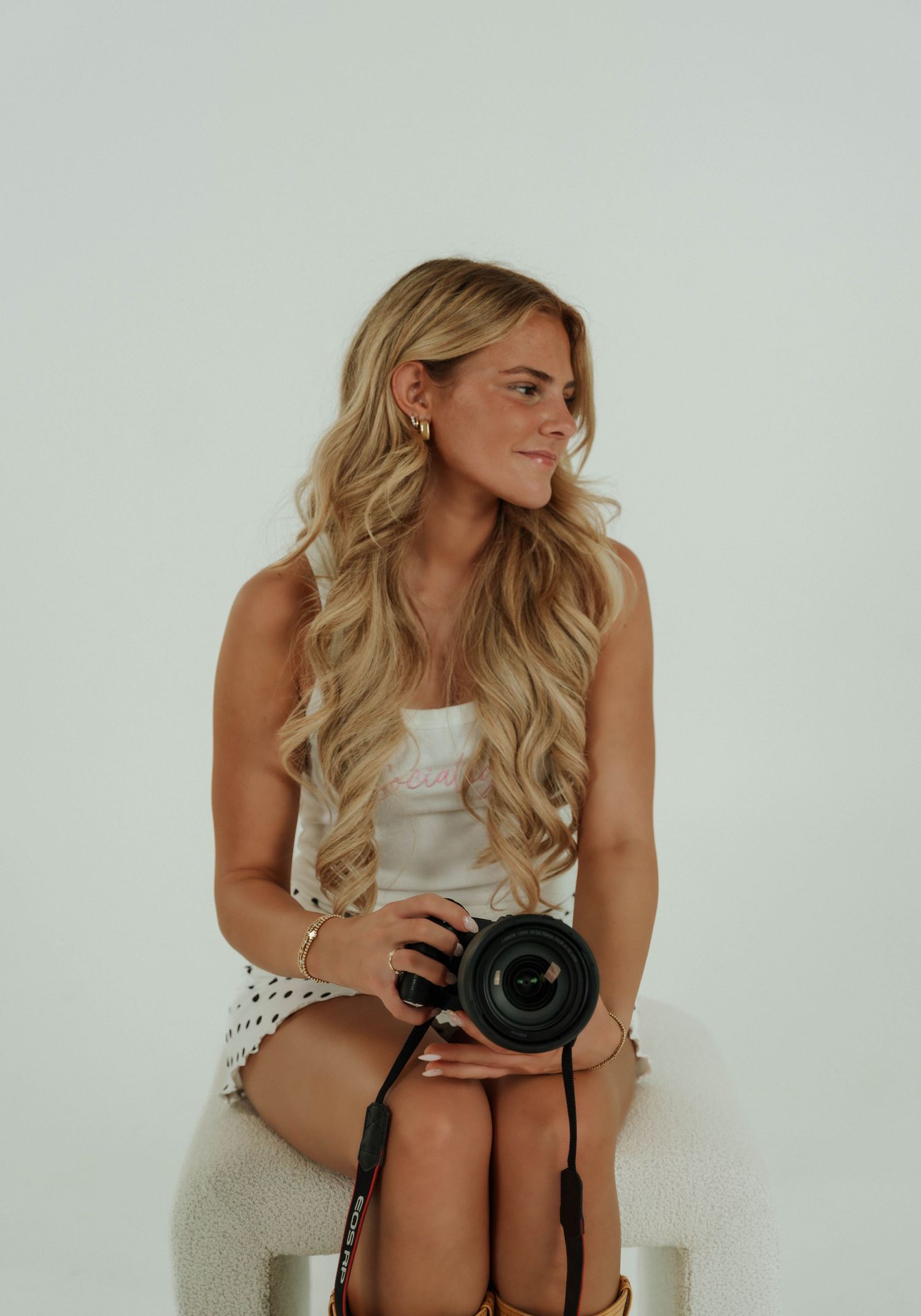 Professional photographer in a white dress sits on a stool while reviewing images on a DSLR camera.