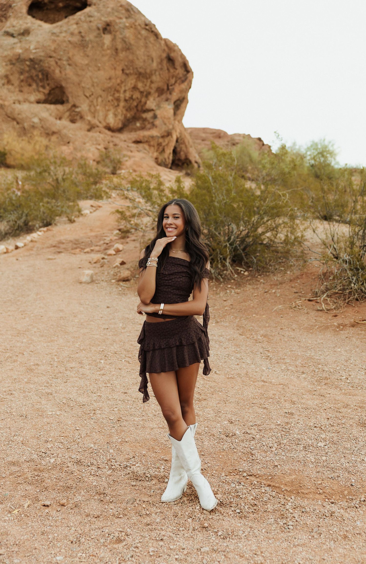 Young woman in brown mini dress and white cowboy boots posing on desert trail with red rock formations.