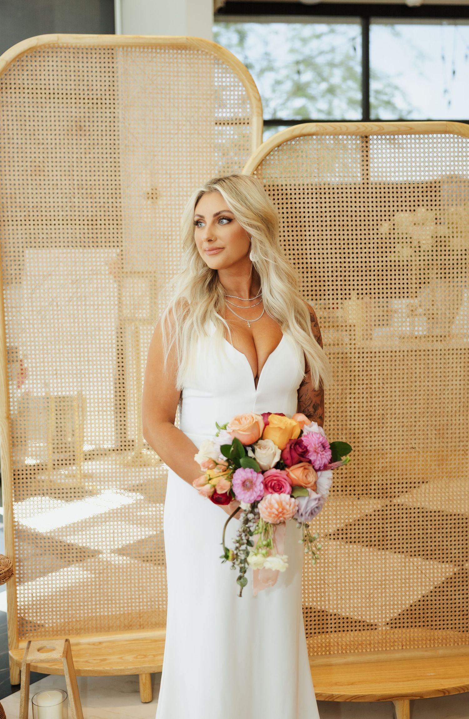 A bride in a white wedding dress holds a vibrant bouquet of pink, orange and peach flowers against a rattan screen backdrop.