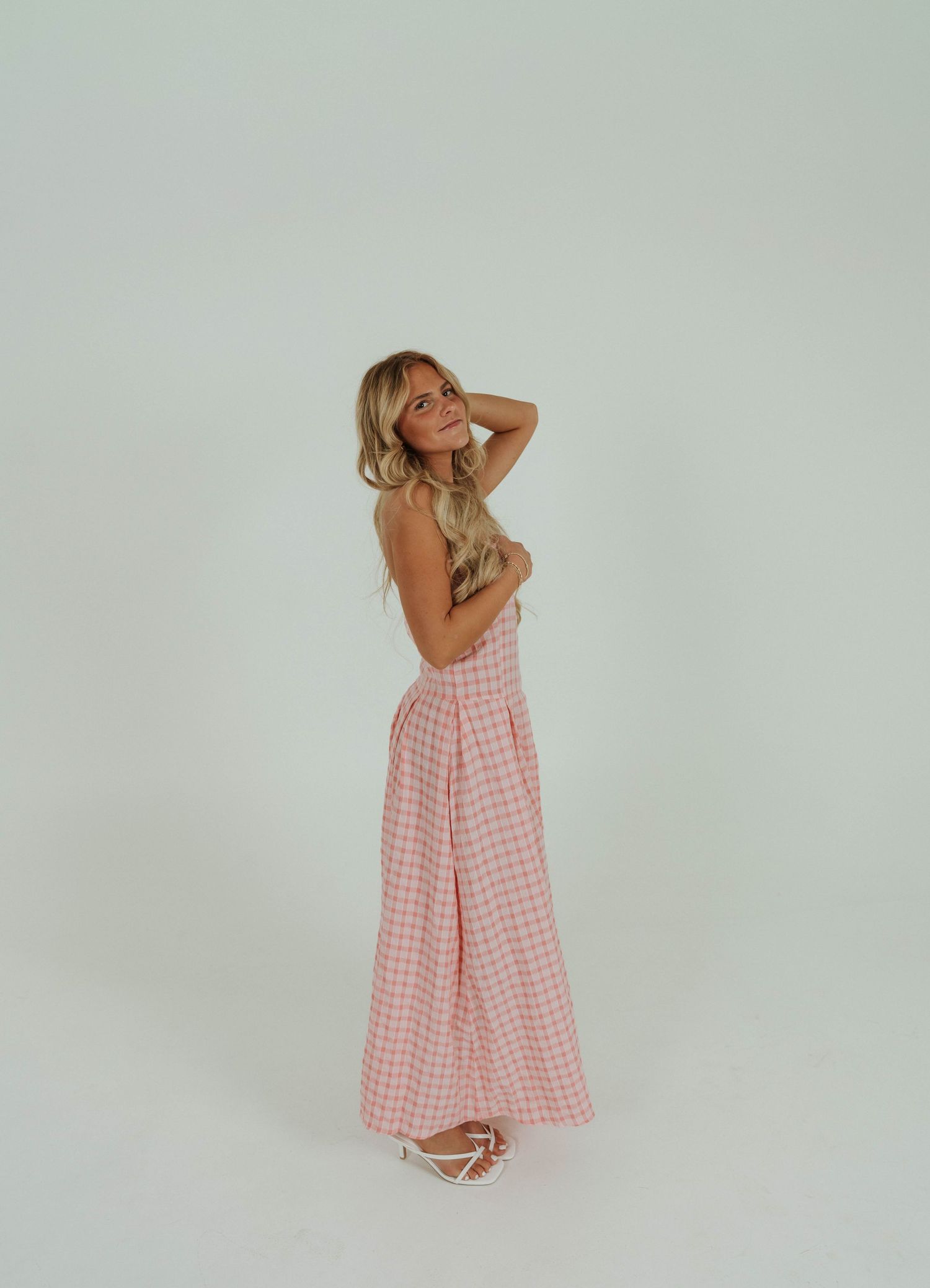 A model in a pink gingham sundress poses playfully in a studio setting against a white backdrop.