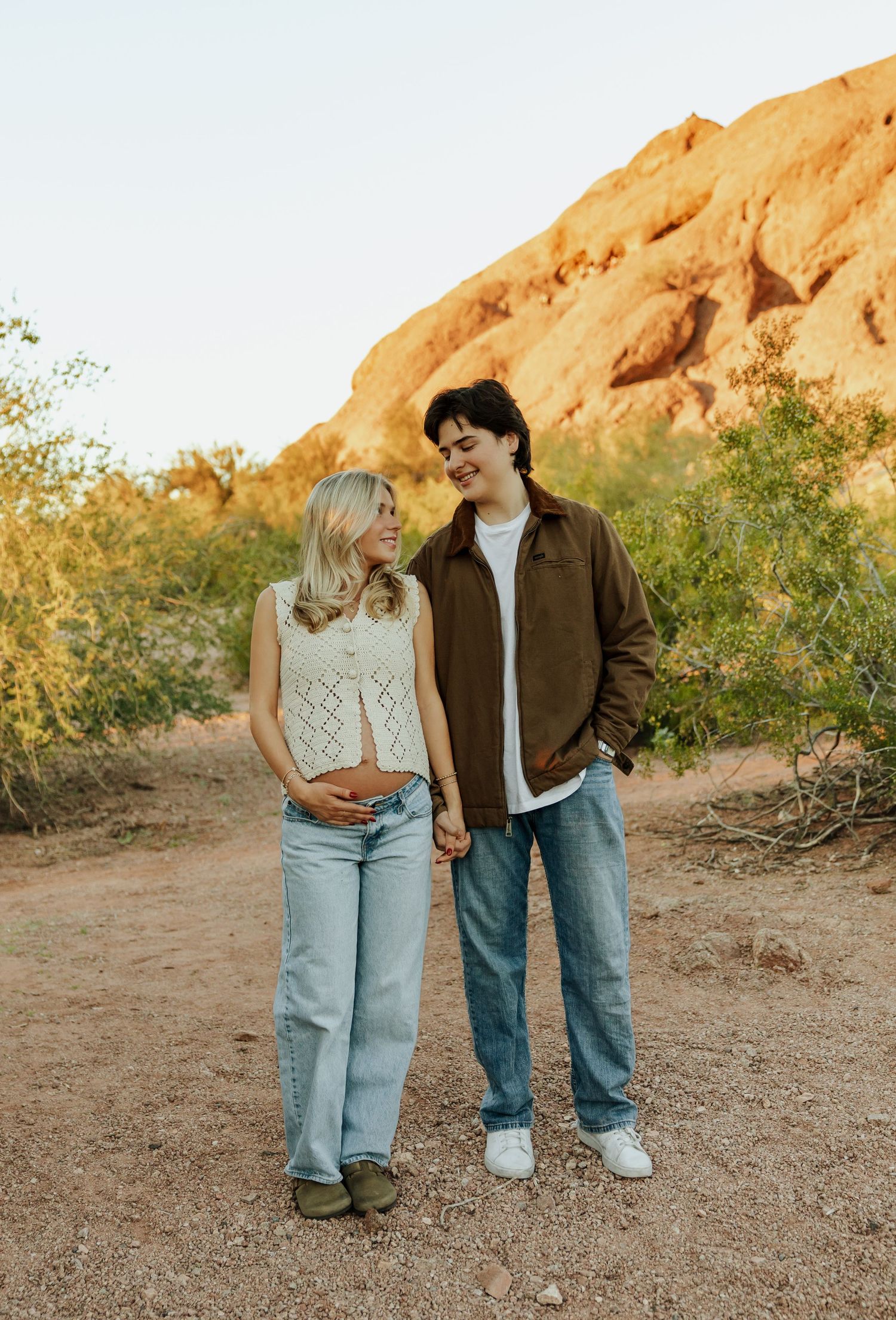 A couple stands together on a dirt path with red rock formations and desert brush in the background at sunset.