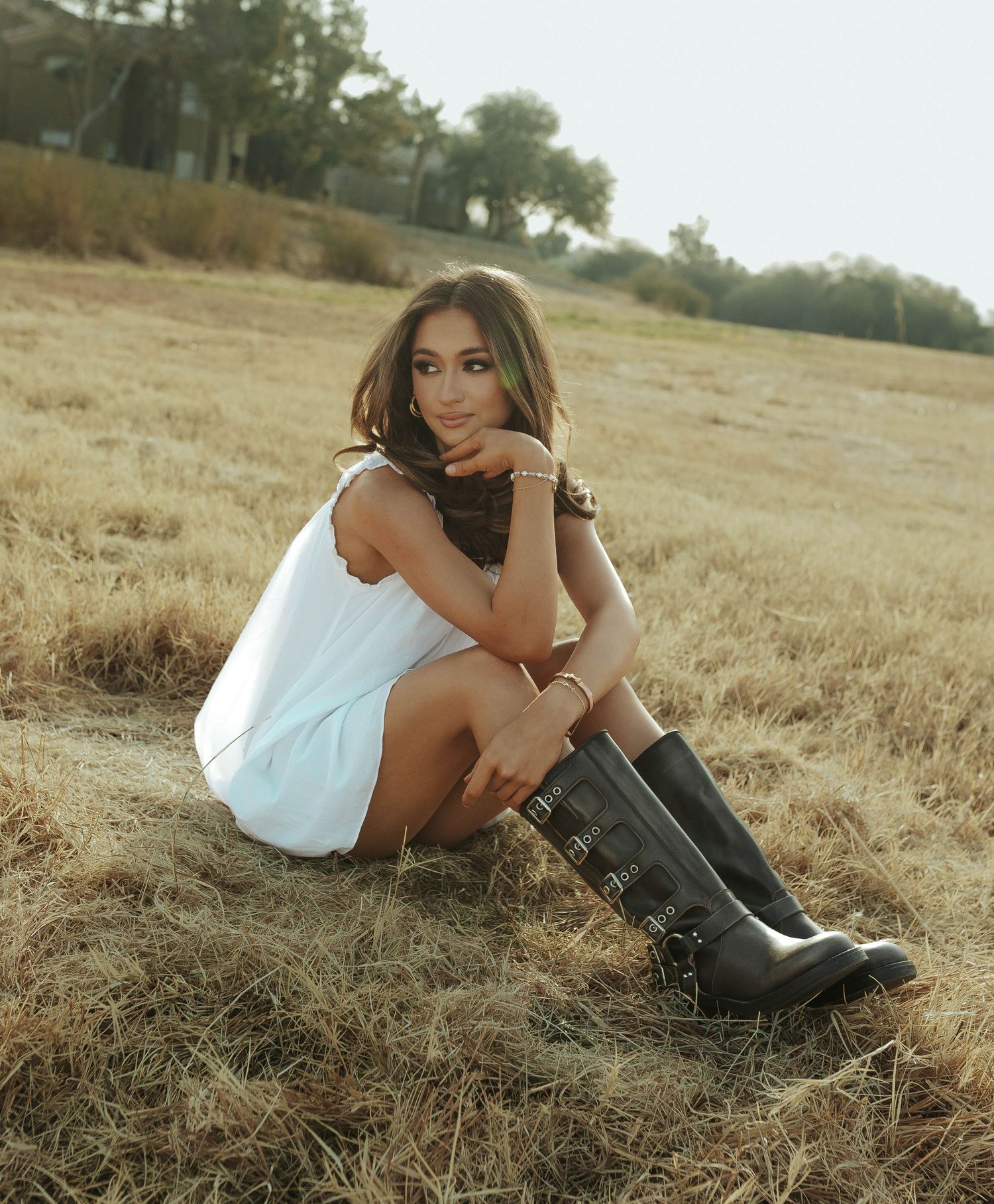 Person wearing a white dress and black boots lies casually in a golden field during a sunny outdoor photoshoot.