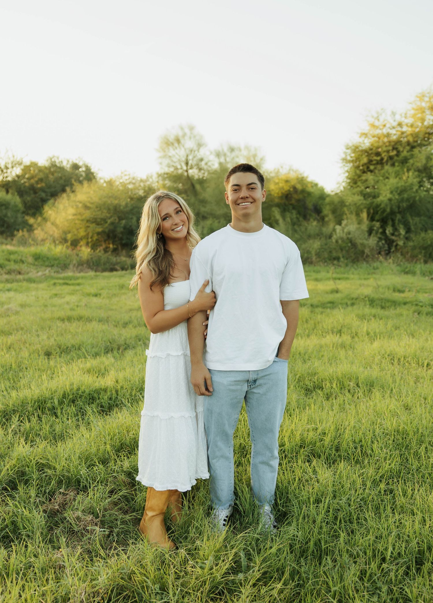A couple stands together in a sunlit meadow wearing white and light blue outfits during a summer photoshoot.