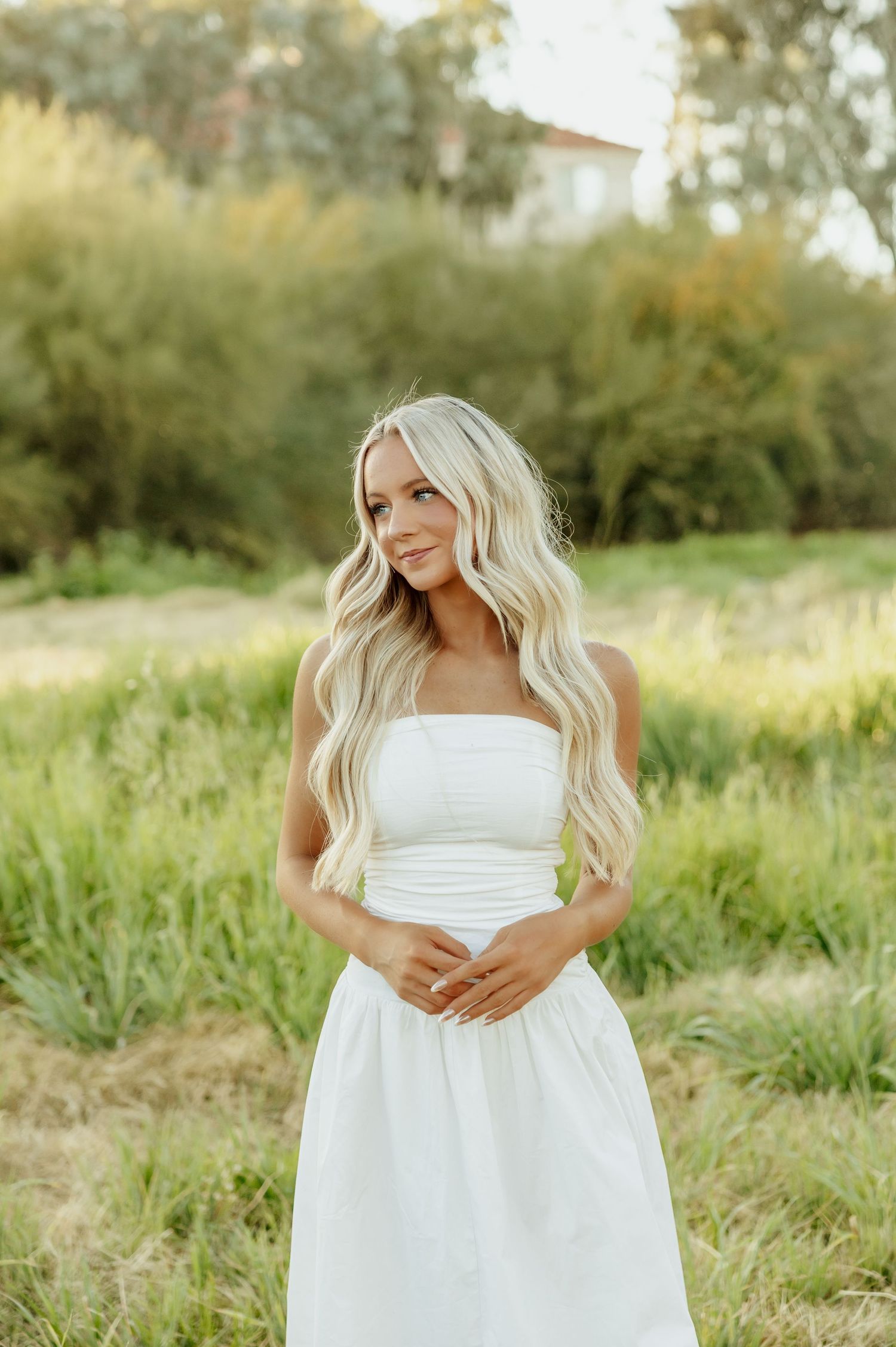 A person in a flowing white dress poses gracefully in a sunlit meadow with tall grass and trees in the background.