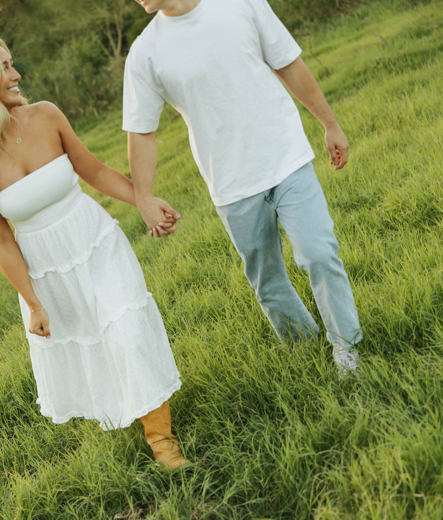 Couple holding hands while walking through lush green grass in white summer attire.