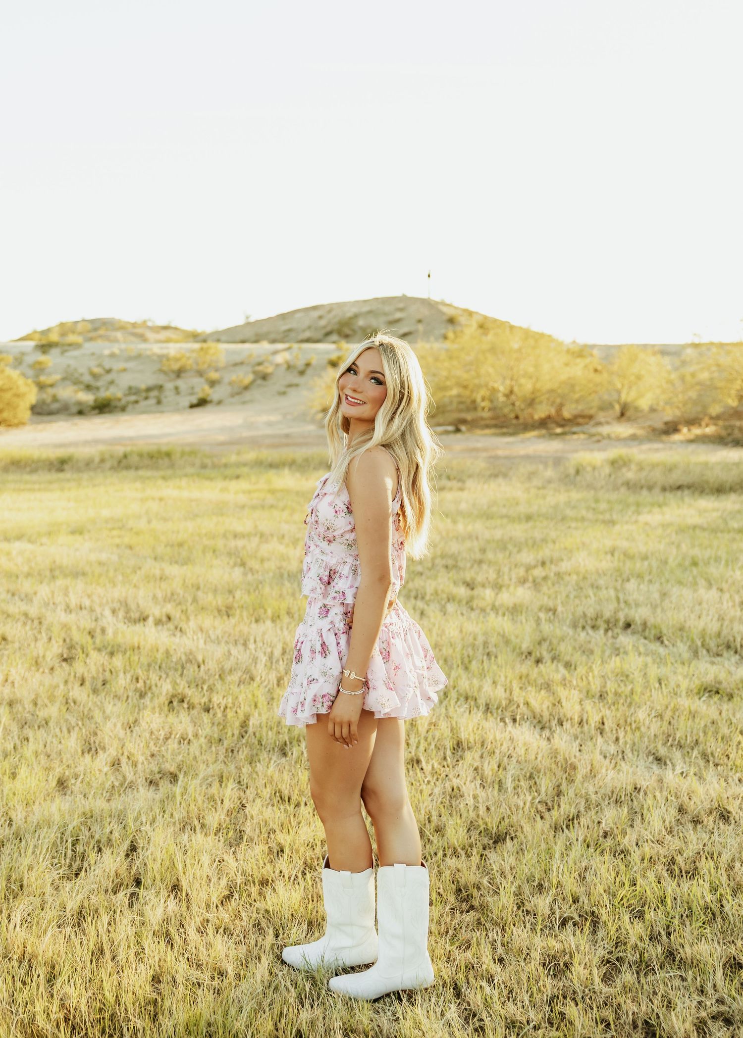 Person in a floral dress and white boots stands in a golden field at sunset.