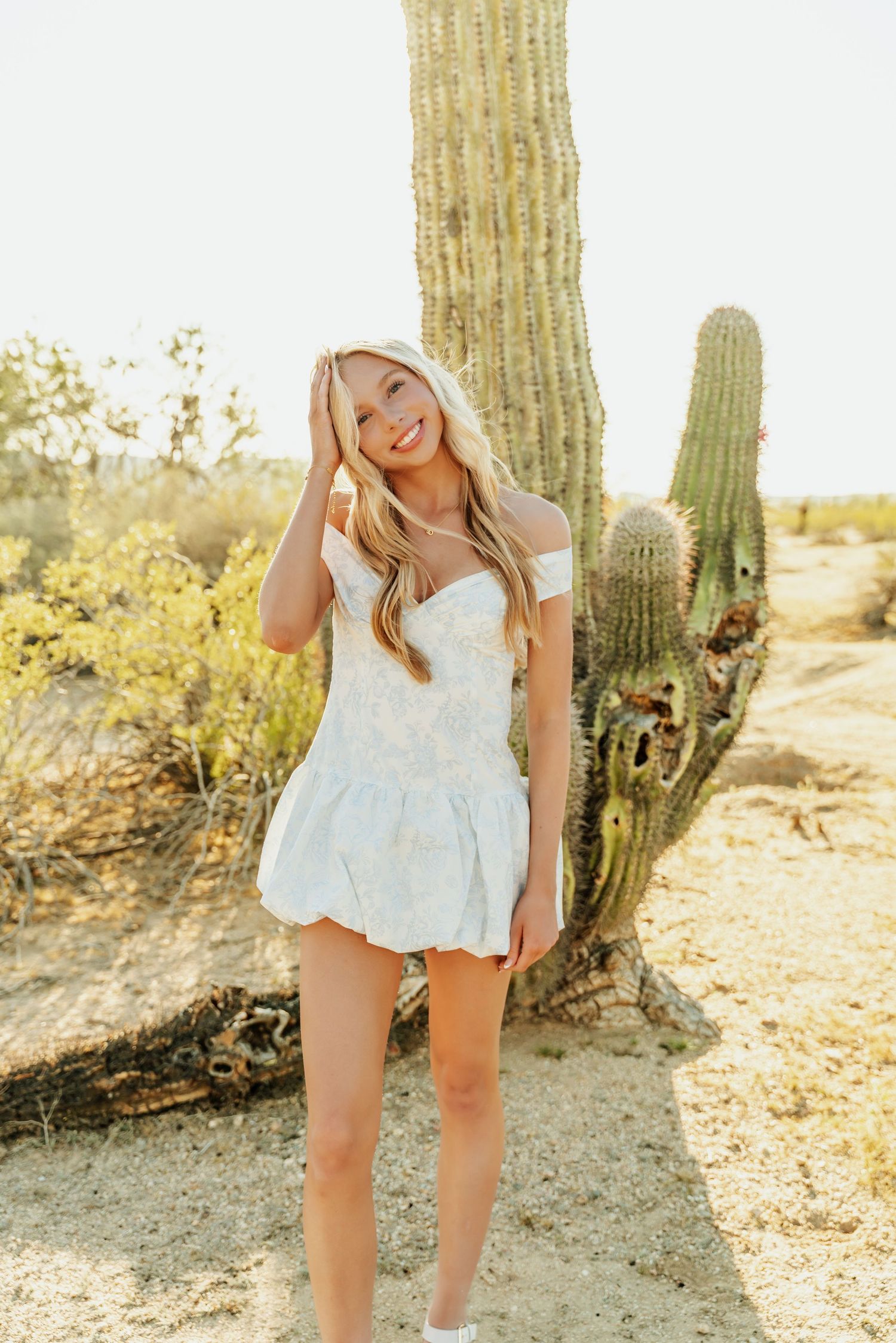 Woman in white summer dress poses next to tall saguaro cactus in sunny desert landscape.