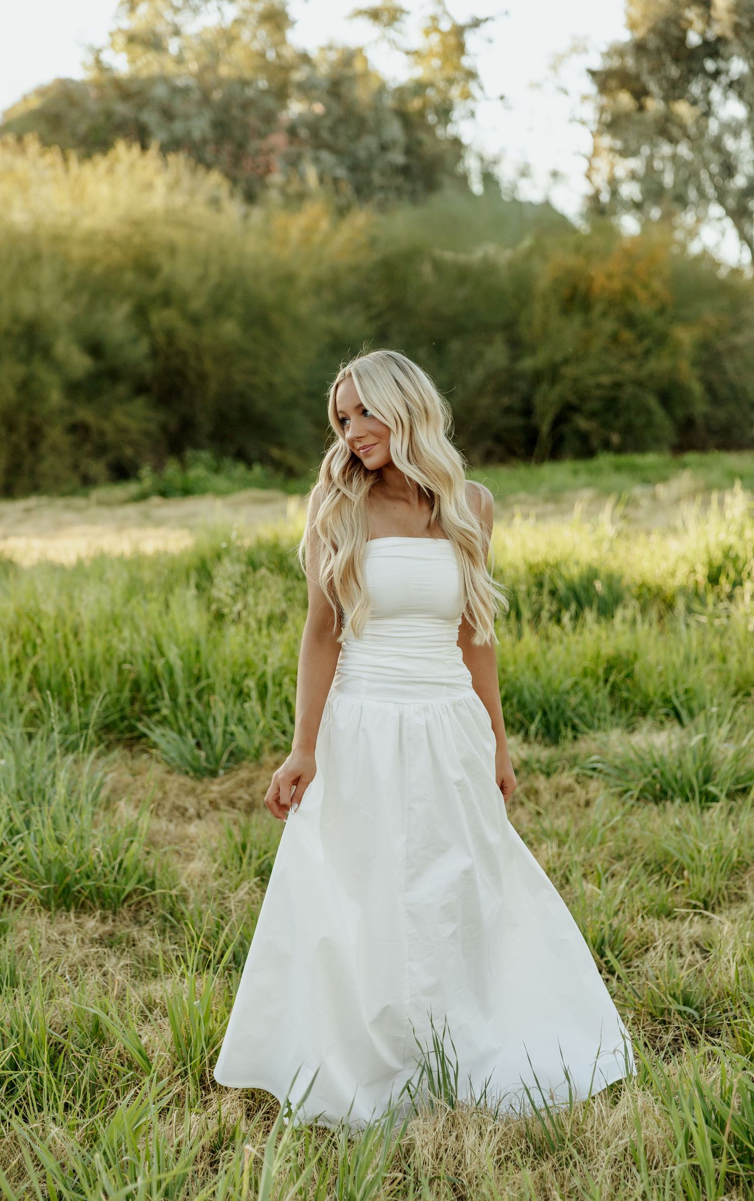 A person in a flowing white wedding gown stands in a sunlit meadow with tall grass and trees in the background.
