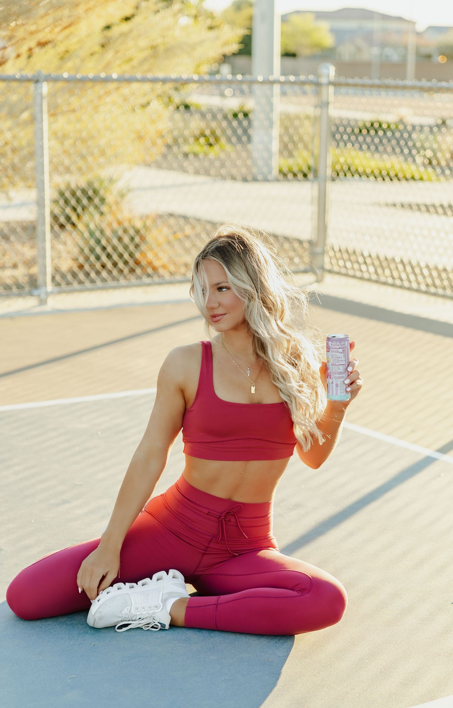 Athletic person in red workout attire sits cross-legged on exercise mat holding energy drink can in outdoor setting.